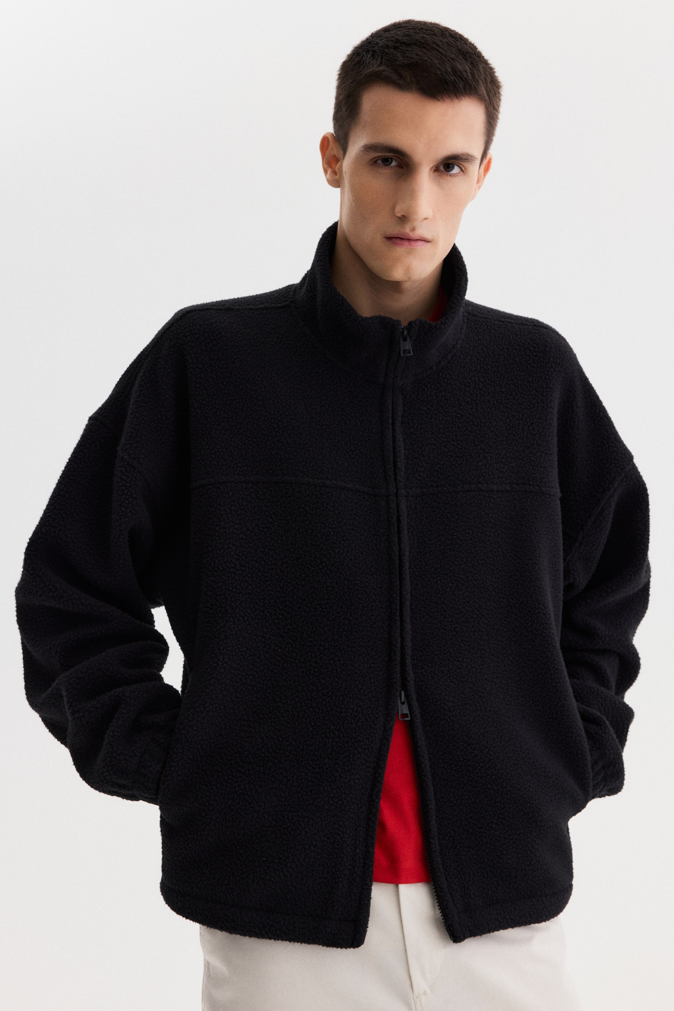View larger image: A black textured fleece jacket faces forward, featuring a full-zip closure and a stand-up collar. It has a relaxed fit with horizontal and vertical seam detailing, partially unzipped over a red shirt and layered over light-colored pants.