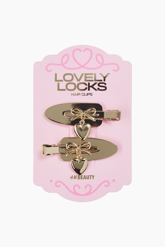 Two gold-toned hair clips are resting facing forwards on a pink display card, with one pointing left and the other pointing right. Each clip features a smooth, oblong metal plate decorated with a small gold bow and a dangling heart charm. - 1