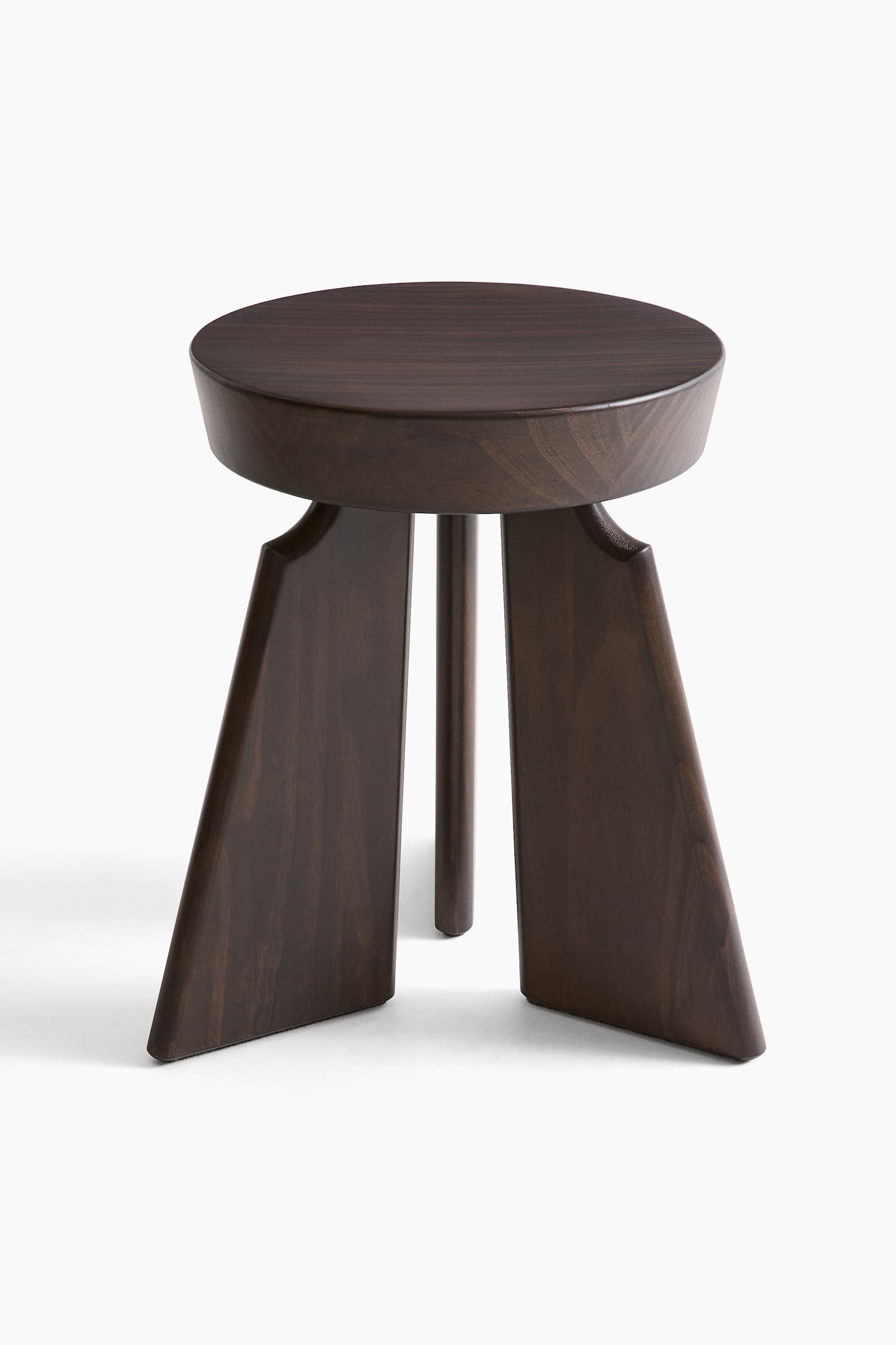 A dark brown wooden stool features a round seat with visible grain, shown from a slight front-right angle, and is supported by three legs. Two wide, flat legs taper downwards and angle outwards from the seat, while one thinner, round central leg is positioned behind them. - 1