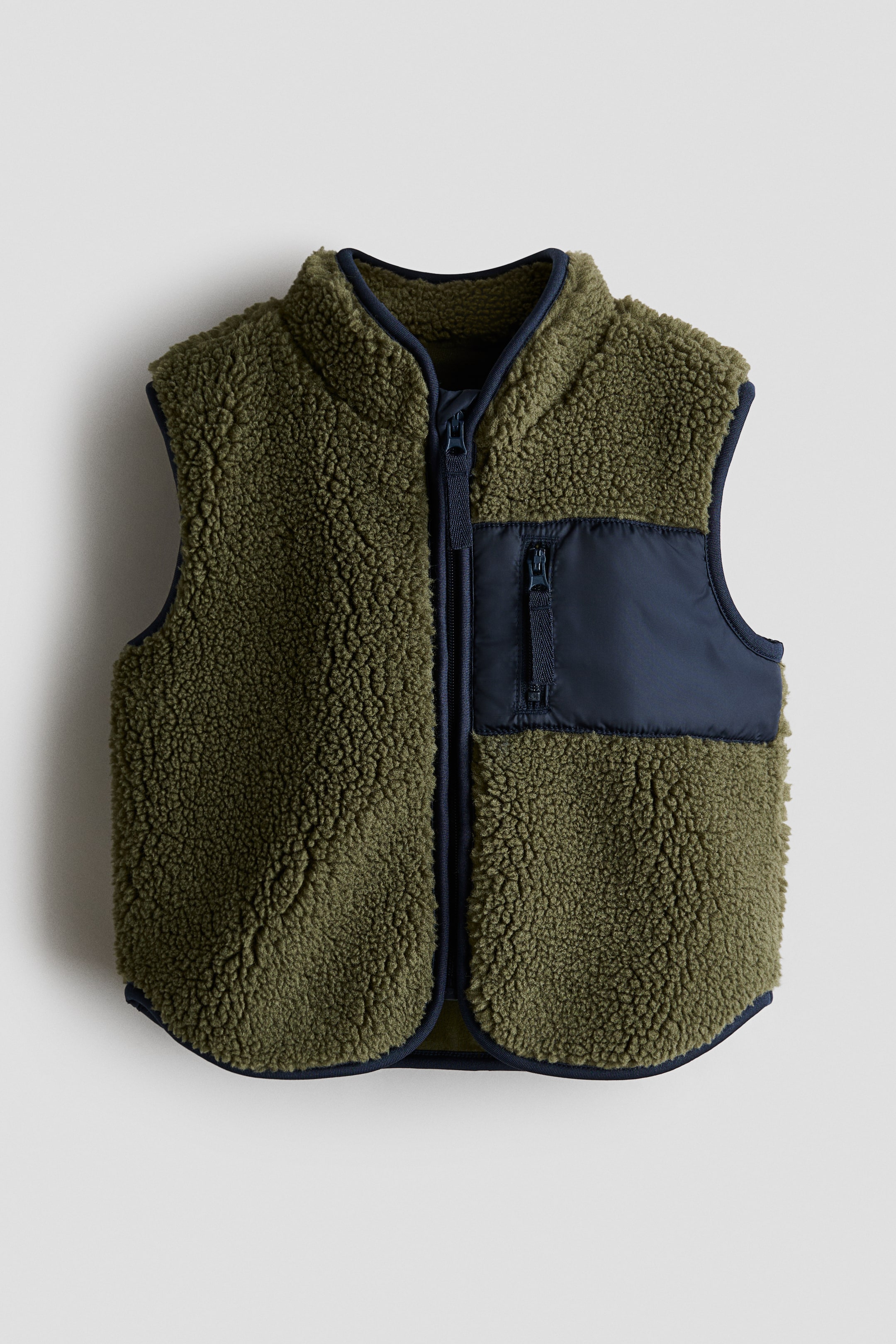 View larger image: The front of an olive green teddy gilet showcases a stand-up collar and a full zip closure with an anti-chafe chin guard. It also features a navy blue zipped pocket crafted from woven fabric on the chest, along with navy jersey bindings that outline the collar, armholes, and hem.