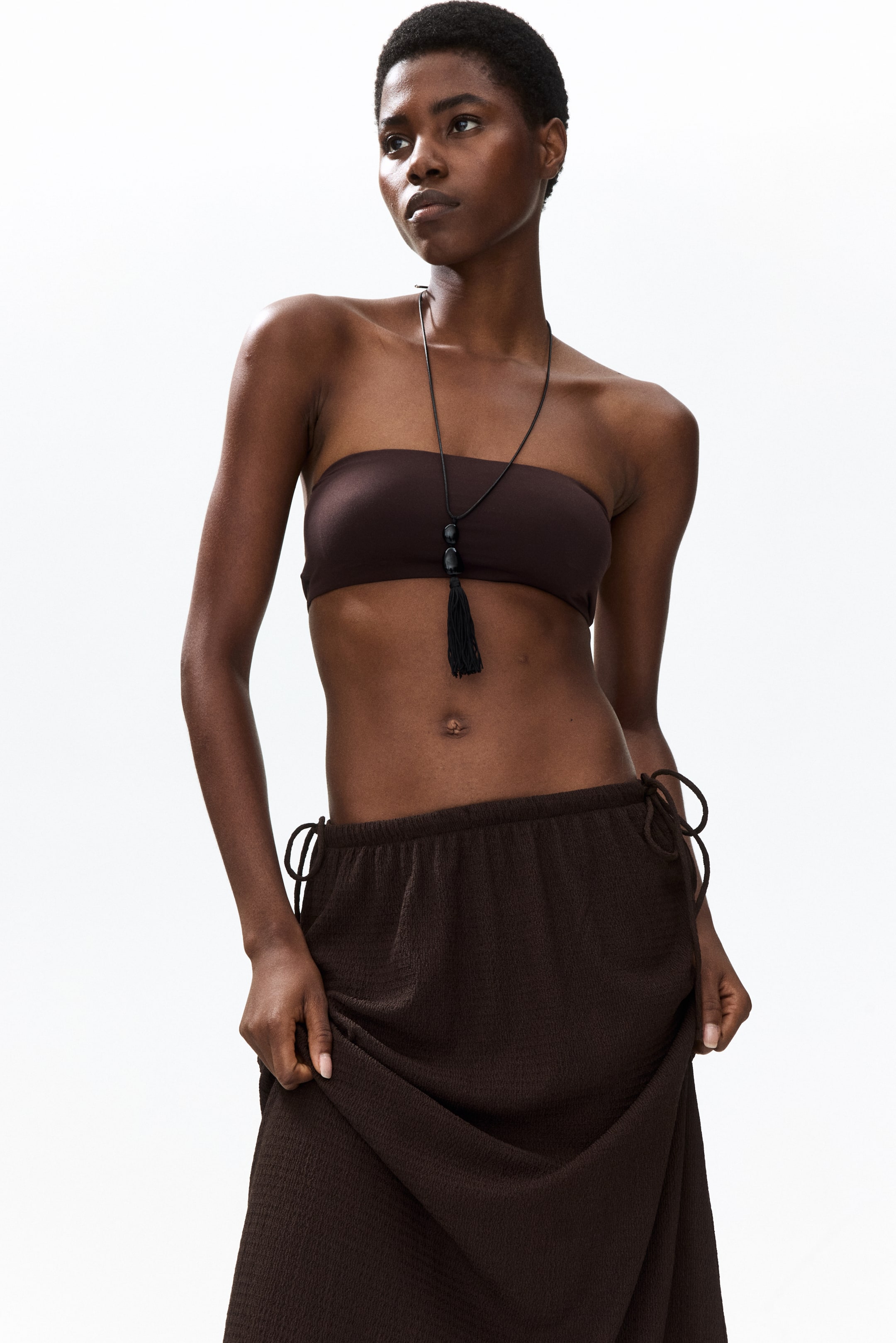 View larger image: A dark brown strapless bandeau top paired with a matching dark brown textured maxi skirt is showcased. The skirt's waist gathers at the sides with visible drawstrings, and its flowing fabric is held up to highlight its drape and texture.