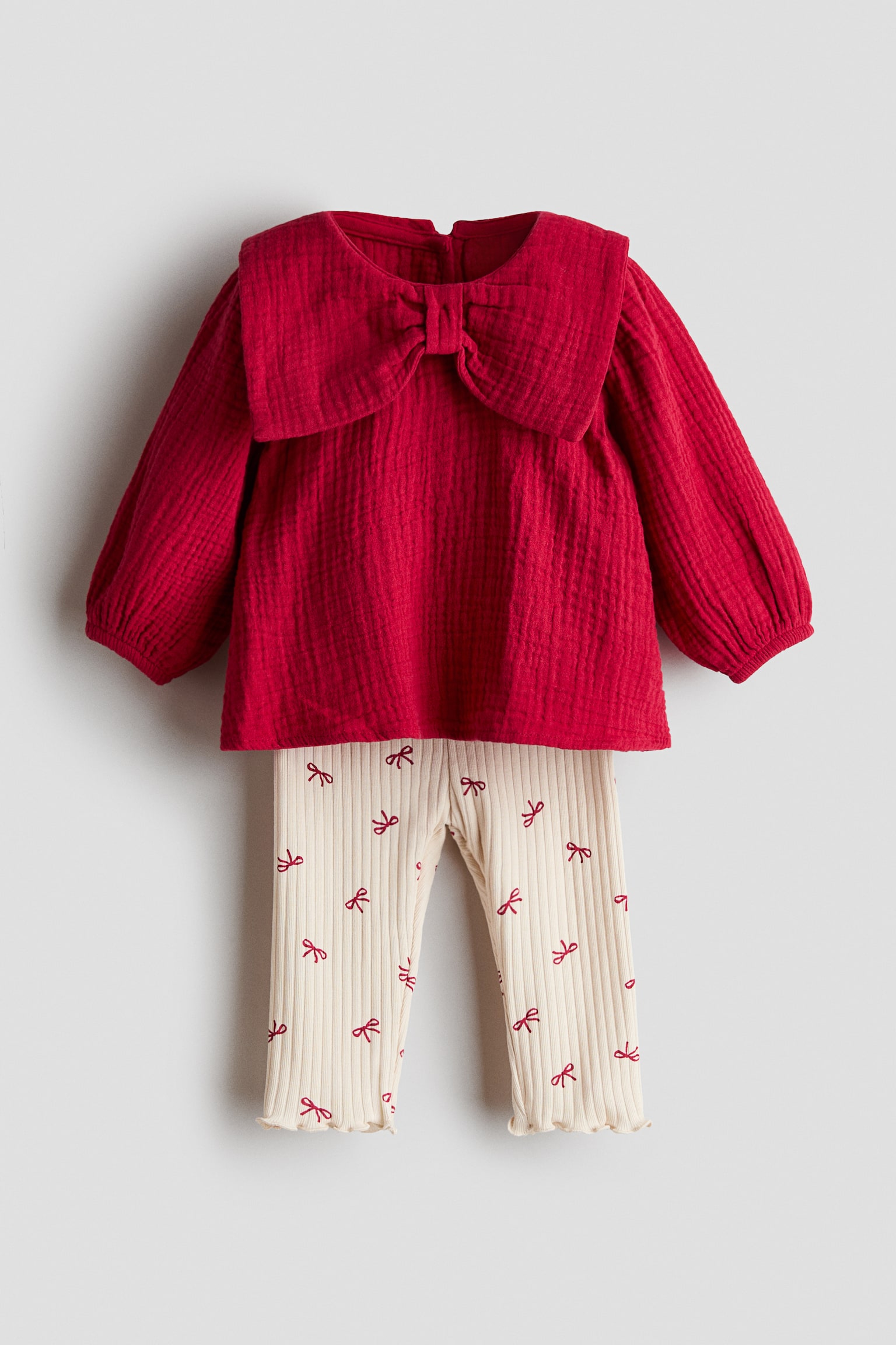 Un ensemble pour enfant composÃ© dâune blouse et dâun legging prÃ©sente une vue de face dâune blouse rouge en mousseline Ã  manches longues et dâun legging cÃ´telÃ© couleur crÃ¨me. La blouse est dotÃ©e dâun grand col oversize en forme de nÅud et de poignets Ã©lastiquÃ©s, tandis que le legging arbore un motif intÃ©gral de nÅuds rouges avec des ourlets surjetÃ©s.