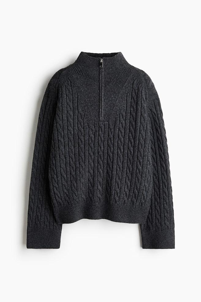Un maglione a trecce grigio scuro è mostrato frontalmente, caratterizzato da un collo alto a coste con chiusura a mezza zip. Il maglione presenta un motivo a trecce verticali su tutta la superficie del corpo e delle maniche lunghe, rifinite con polsini a coste e un orlo a coste. - 1