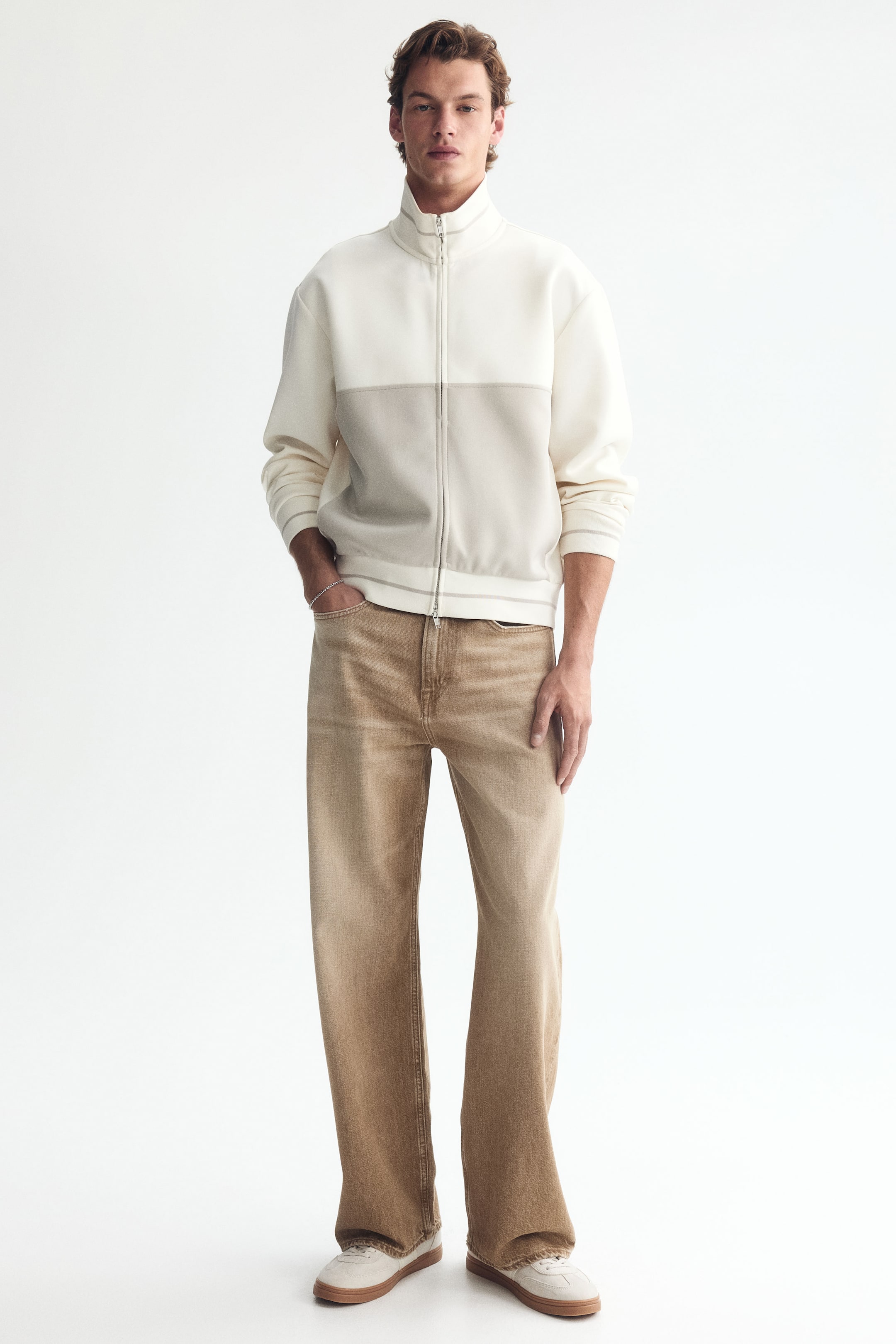View larger image: The model is donning light brown 5-pocket jeans crafted from sturdy cotton denim. These jeans feature a regular waist, straight leg, and offer a relaxed fit from the seat to the hem. They have a dropped crotch and provide ample room throughout the leg. The model is posed with one hand casually placed in a front pocket of the jeans.