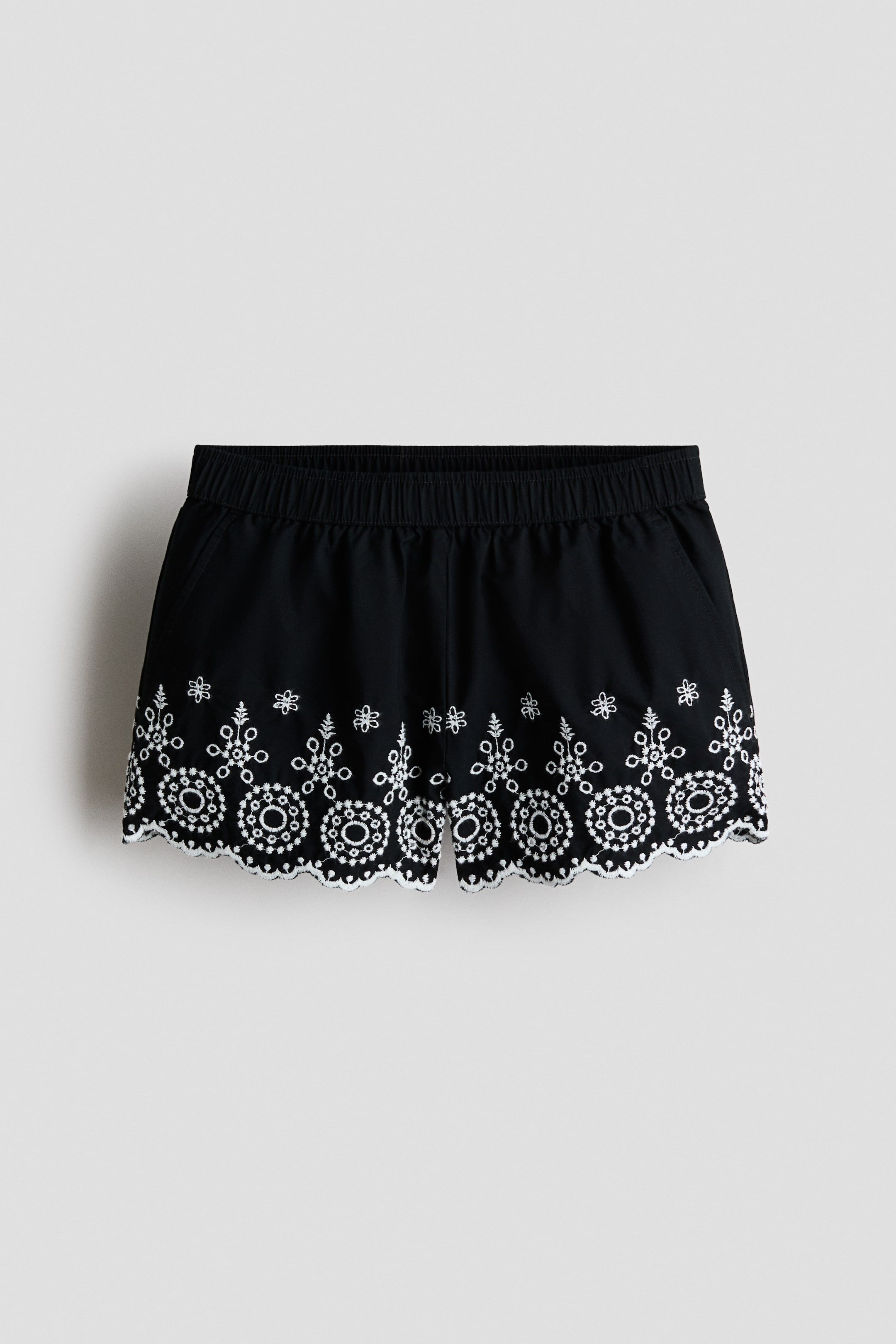 View larger image: Black shorts feature an elastic waistband and side pockets. They showcase white embroidered eyelet detailing with a scalloped hem along the bottom edge.