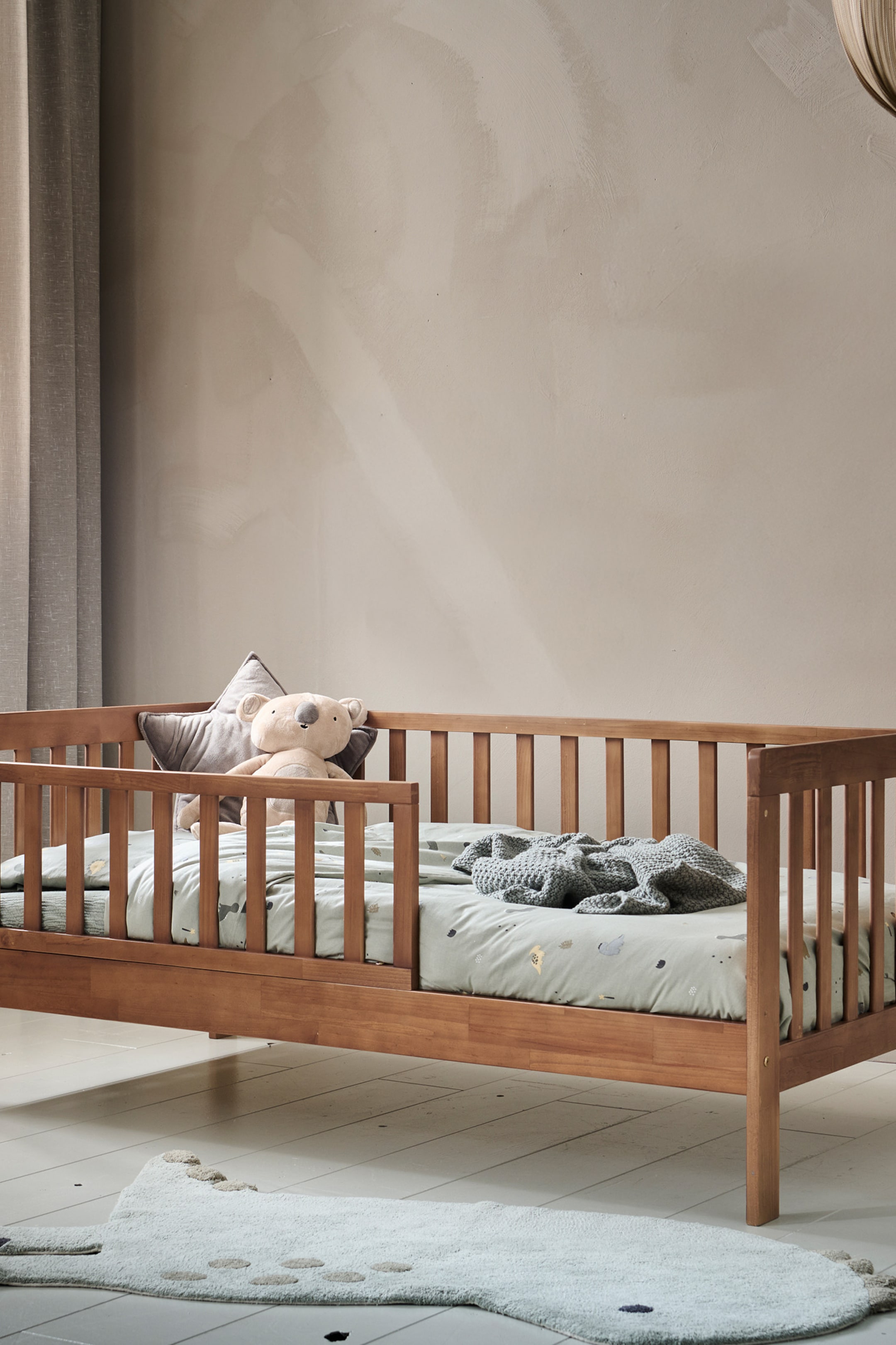 Agrandir l'image: Un lit pour tout-petit en bois est légèrement incliné vers la droite, avec un design bas et des barrières latérales à lattes. Il présente un drap vert clair avec des motifs subtils, une couverture grise en tricot, un coussin en forme d'étoile gris, et un jouet en peluche koala reposant sur le matelas.