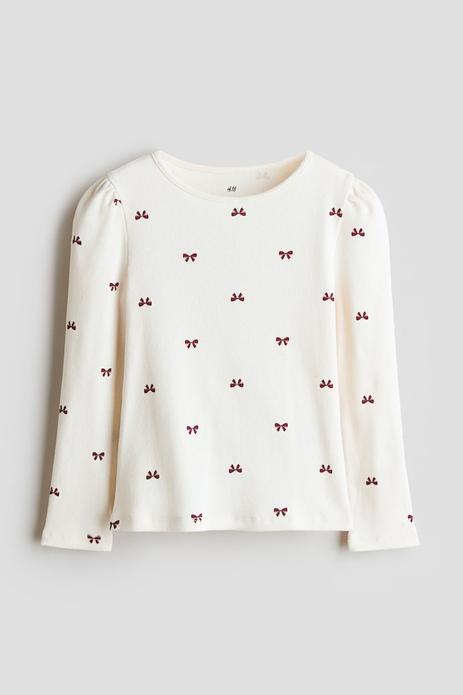 Un haut de couleur crème en jersey de coton côtelé est présenté de face, avec un col rond et des manches longues bouffantes à épaules froncées. Le haut est orné dâun motif all-over de petits nÅuds bordeaux foncé. - 1