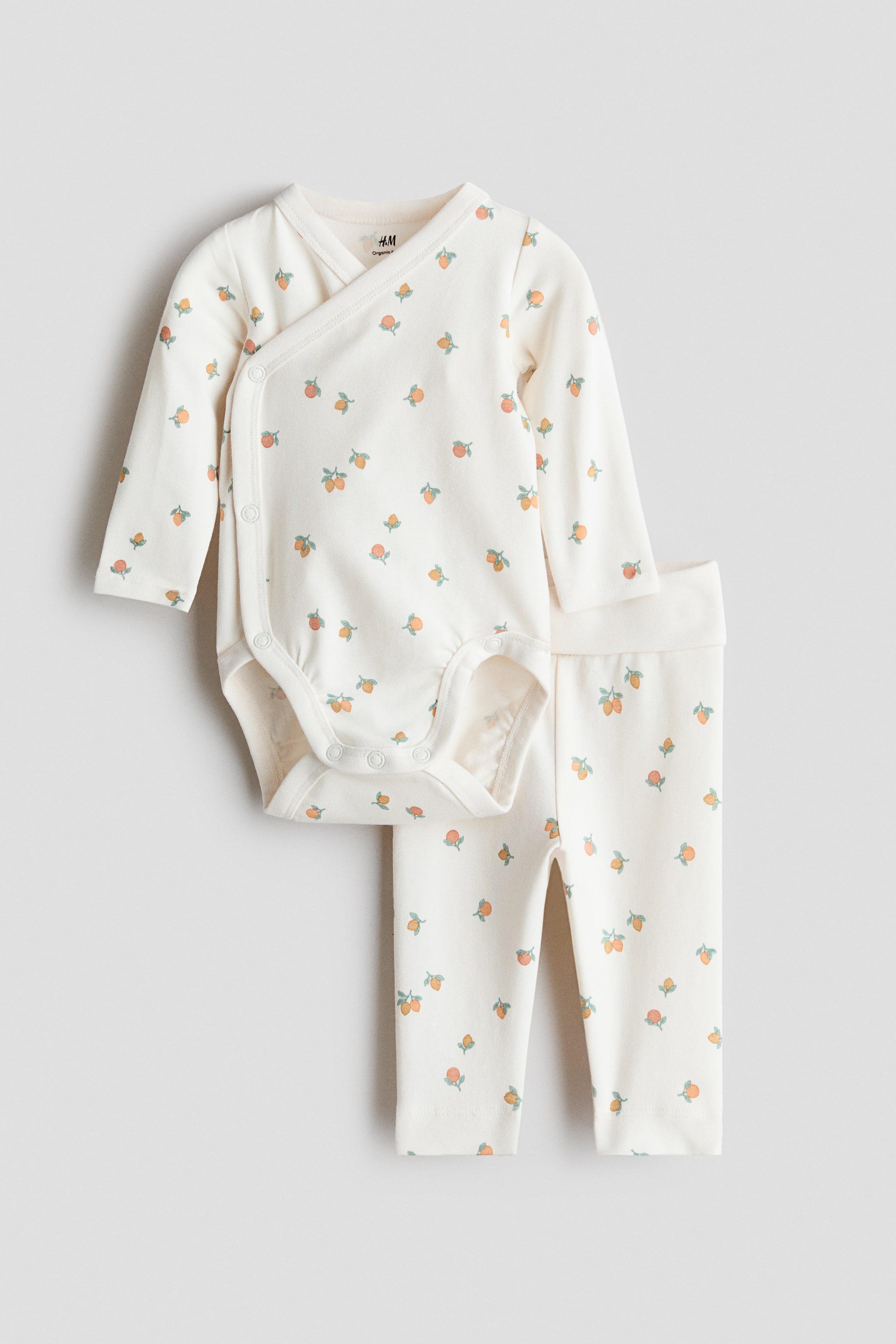 View larger image: A set comprising a bodysuit and leggings is presented in a cream shade with a pattern of small orange and green fruit. The long-sleeved wrapover bodysuit features snap buttons at the side and crotch, while the leggings come with a foldover waist.