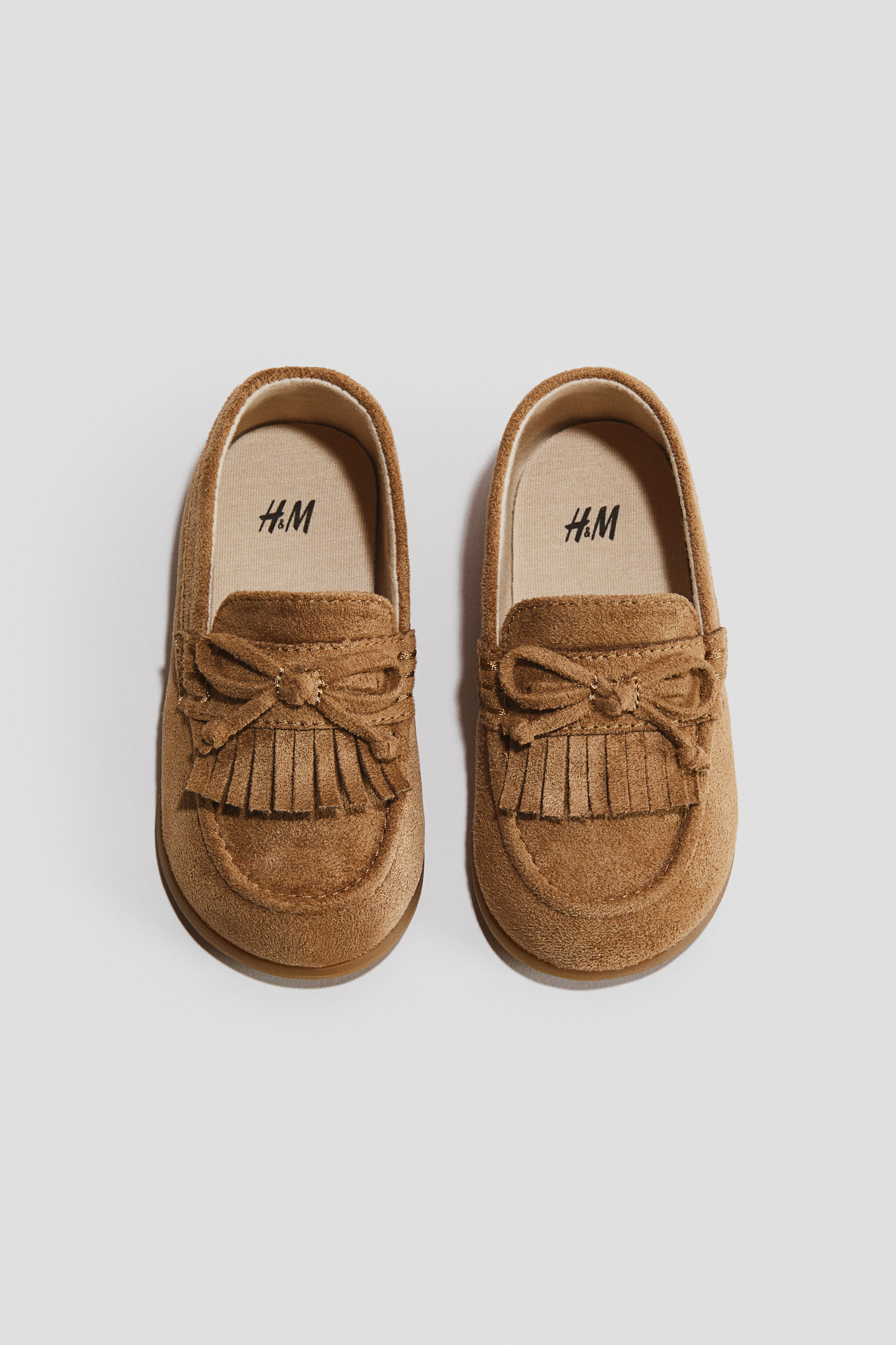Vezi imaginea mai mare: Loafers fără căptușeală sunt prezentate ca o pereche, orientate spre față. Acestea sunt realizate dintr-un material asemănător cu pielea întoarsă de culoare cafenie, cu cusături în stil mocasin în jurul vârfului și un fundiță decorativă cu detalii de franjuri pe vamp; logo-ul H&M apare pe tălpile interioare de culoare bej deschis.