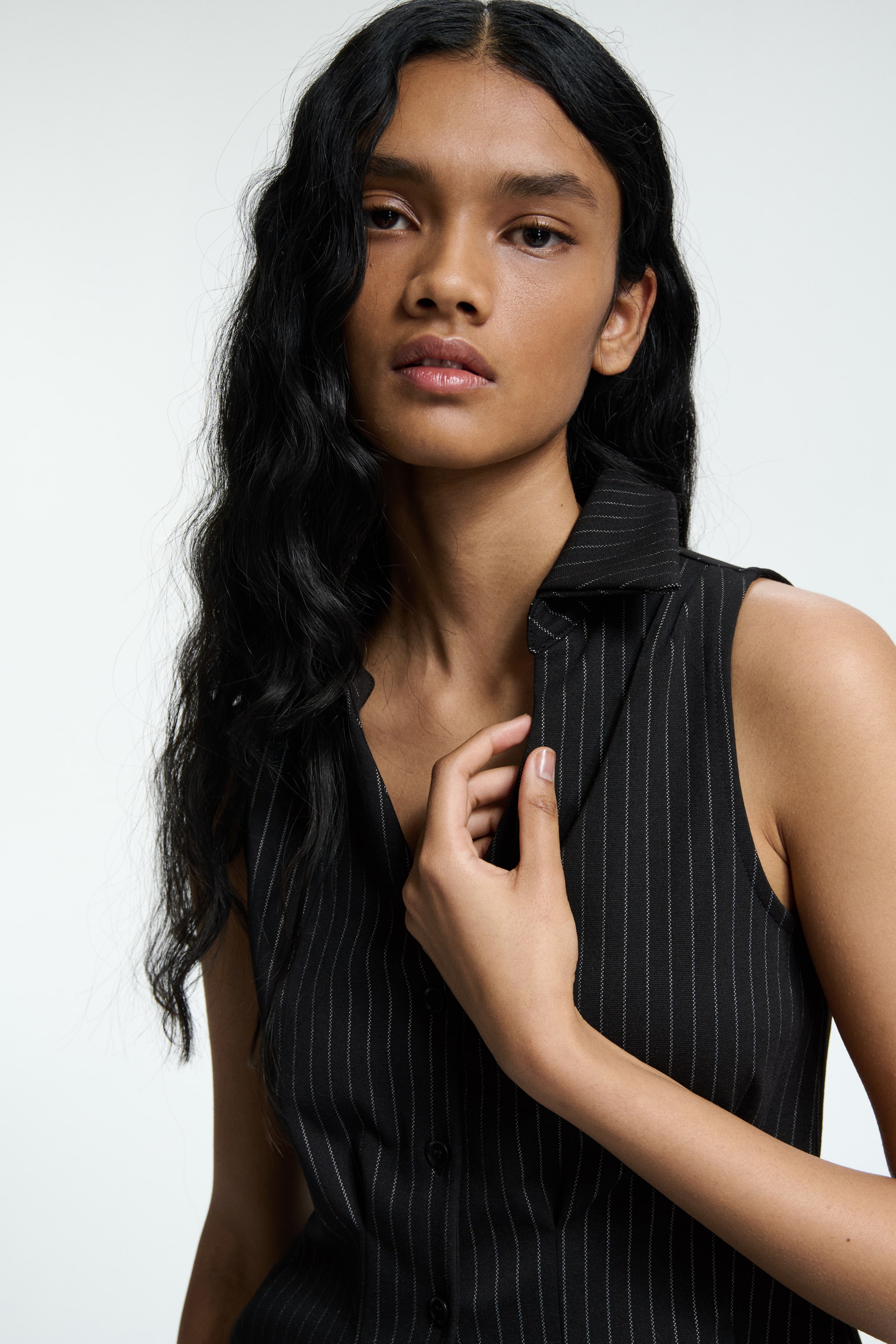 View larger image: A black pinstripe sleeveless vest is displaying a pointed collar and a front button closure. A hand is adjusting the collar, while fine white vertical stripes are running across the fabric.