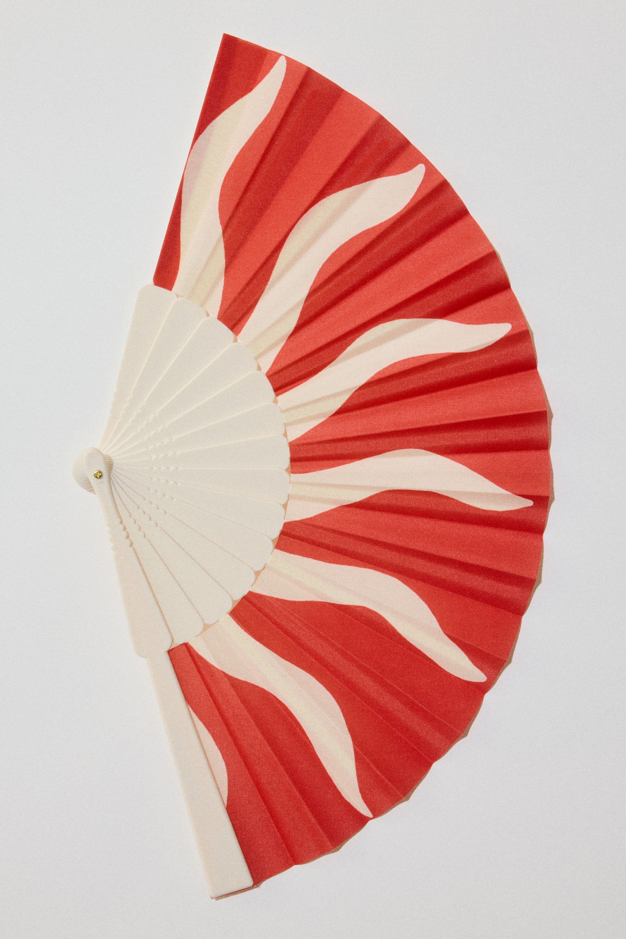 Ver imagen más grande: Un abanico plegable está inclinado desde la izquierda, abriéndose para revelar sus varillas de color claro y su tela plisada. La tela presenta una base roja vibrante con patrones ondulados de color crema que se irradian hacia afuera.
