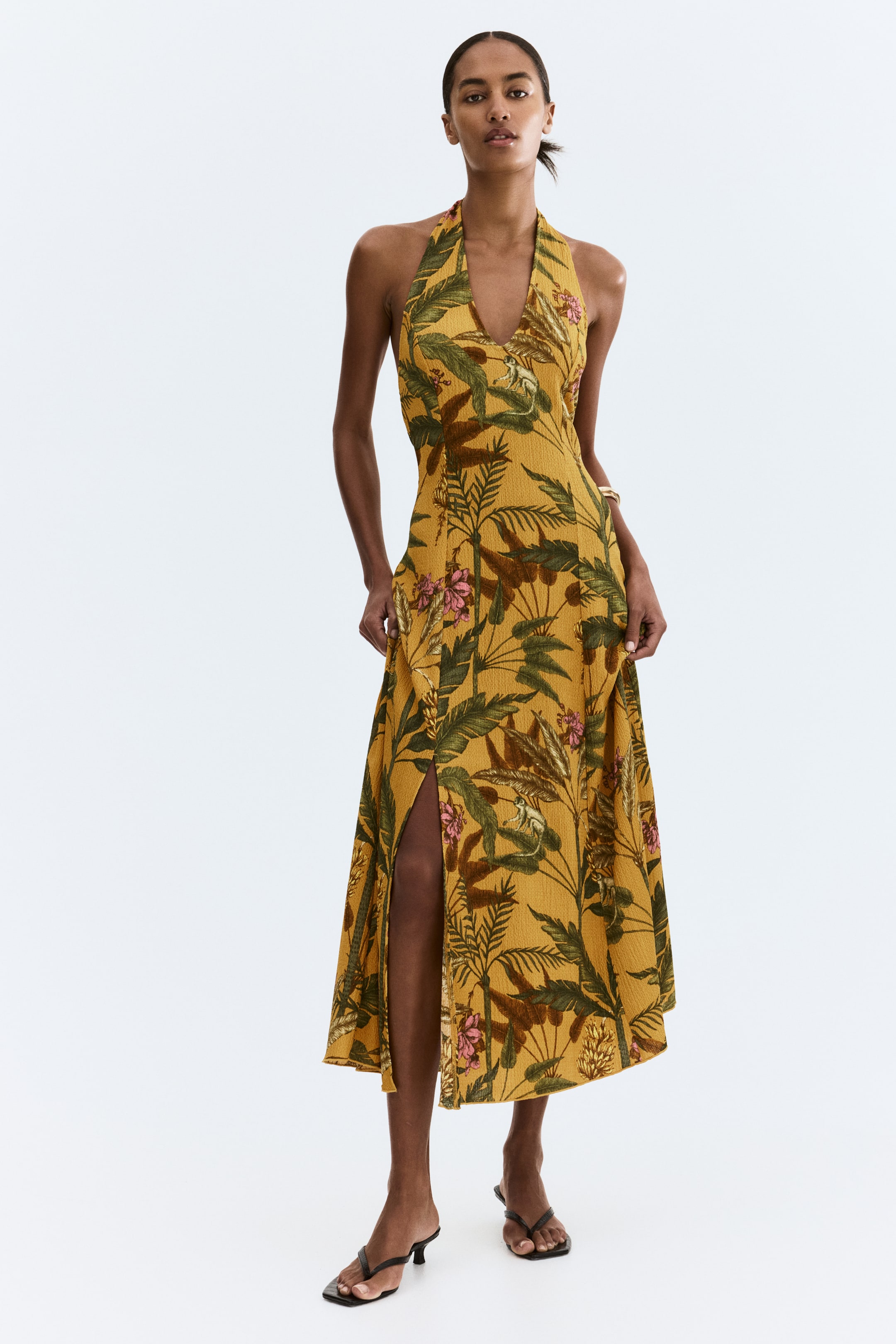 Ampliar la imagen: Un vestido midi amarillo mostaza presenta un estampado tropical con hojas verdes, flores rosas y monos. Está diseñado con un escote halter, un escote en V y una falda fluida con una abertura alta en el costado.