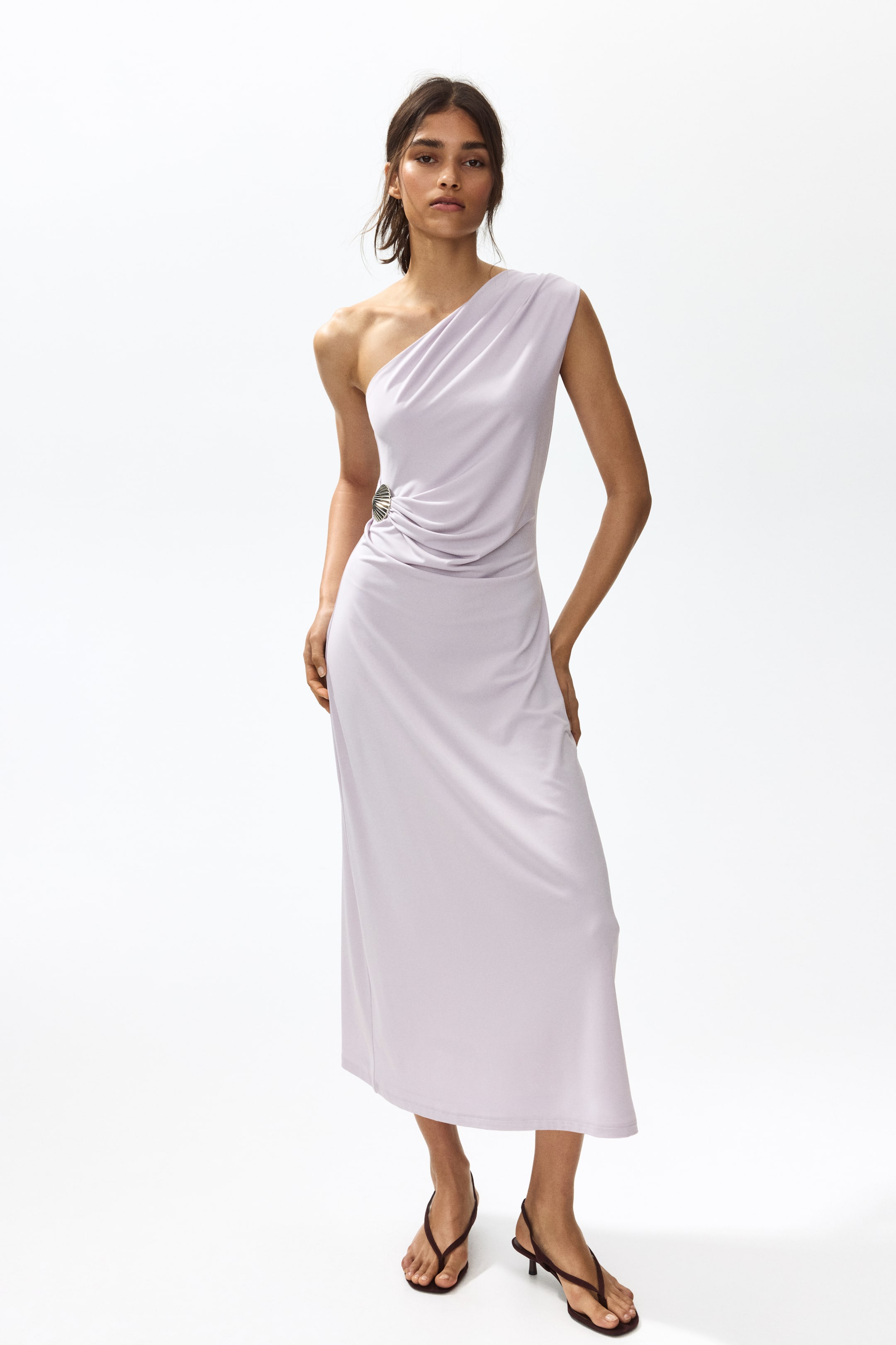 Agrandir l'image: La mannequin porte une robe violette claire, longueur mollet, à une épaule en jersey stretch. La robe ajustée est doublée en haut et présente une large bretelle plissée sur l'épaule gauche de la mannequin, avec une décoration en métal en forme de coquillage à la taille gauche de la mannequin et des plis créant un effet drapé délicat en dessous.