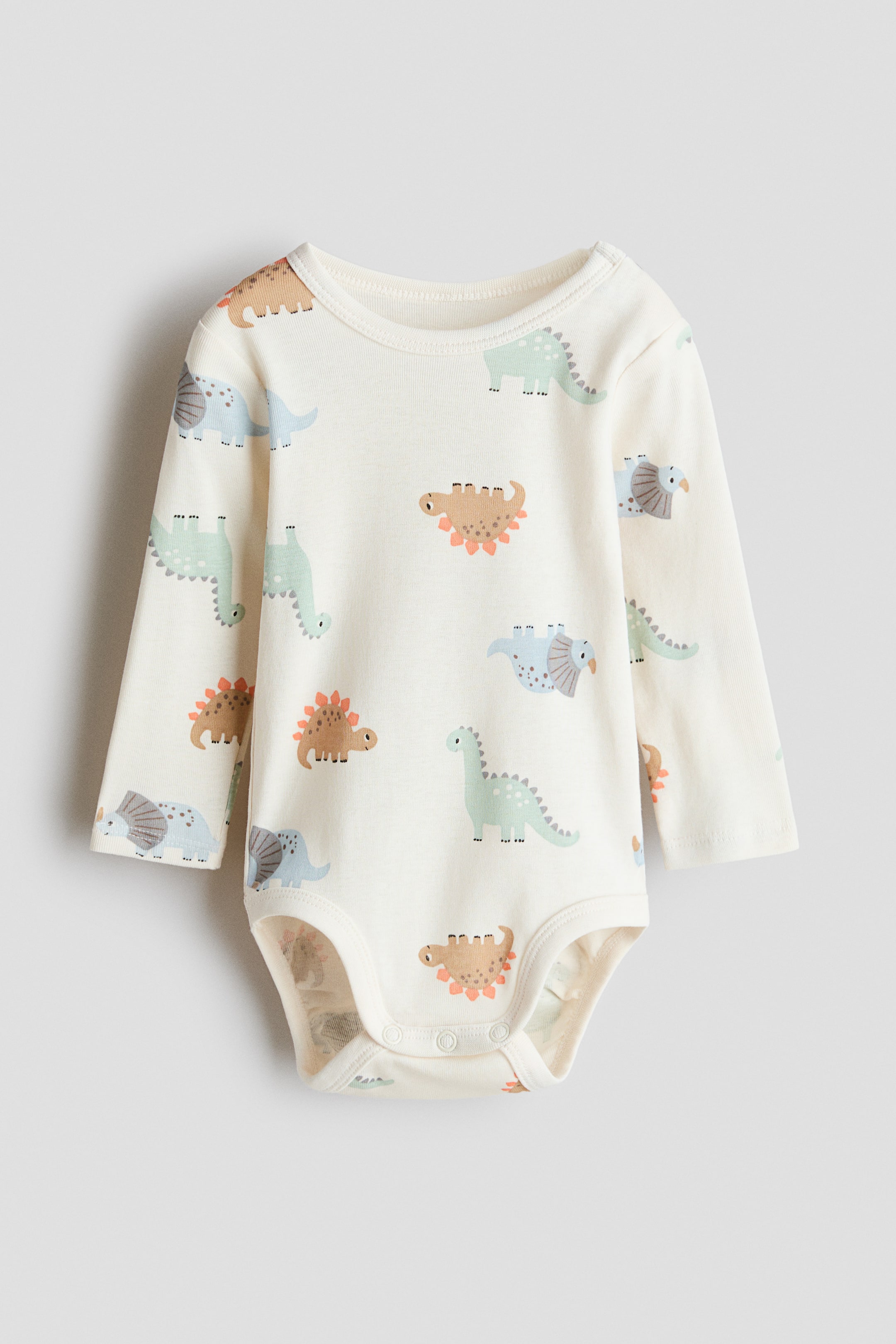 Näytä suurempi kuva: Pehmeästä puuvillatrikoosta valmistettu kermavärinen body esitellään EDESTÄ, ja siinä on kuvio eri sarjakuvadinosauruksista sinisenä, vihreänä ja ruskeana. Se sisältää pyöreän, kantatun kaula-aukon, pitkät hihat, nepparin vasemmalla olkapäällä ja piilotetut nepparit alaosassa.