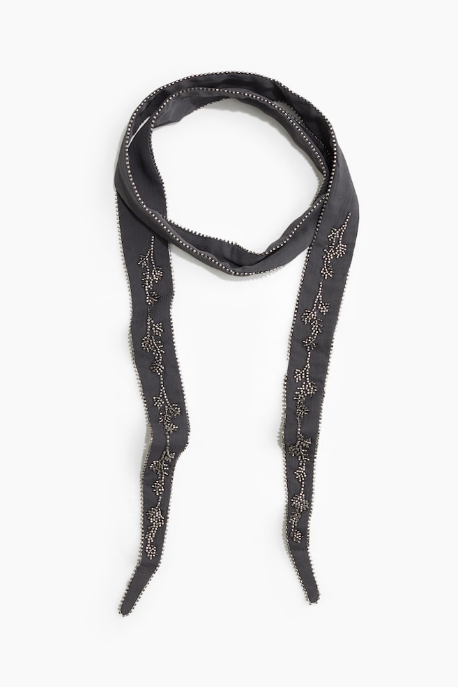 A long, narrow dark gray chiffon scarf is lying flat from the front, arranged in a loose loop with two ends hanging down. Small silver-toned beads are lining the edges and forming intricate, branch-like decorative patterns along the lower halves of both ends. - 1