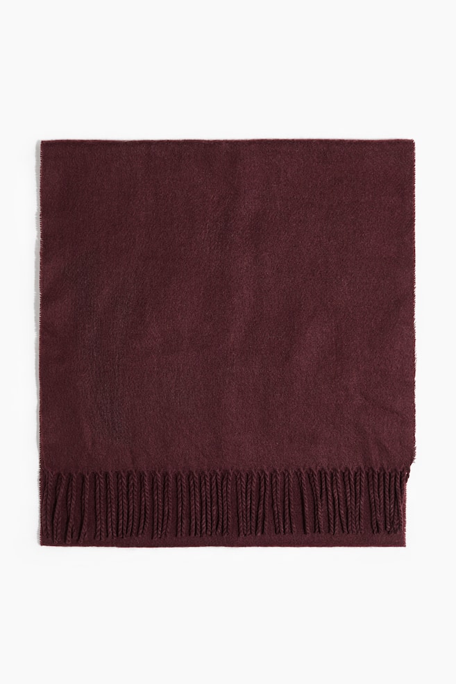 Um cachecol vermelho-escuro, feito de tecido tecido com um acabamento suave e escovado, está deitado plano, exibindo franjas ao longo dos seus lados curtos. - 1