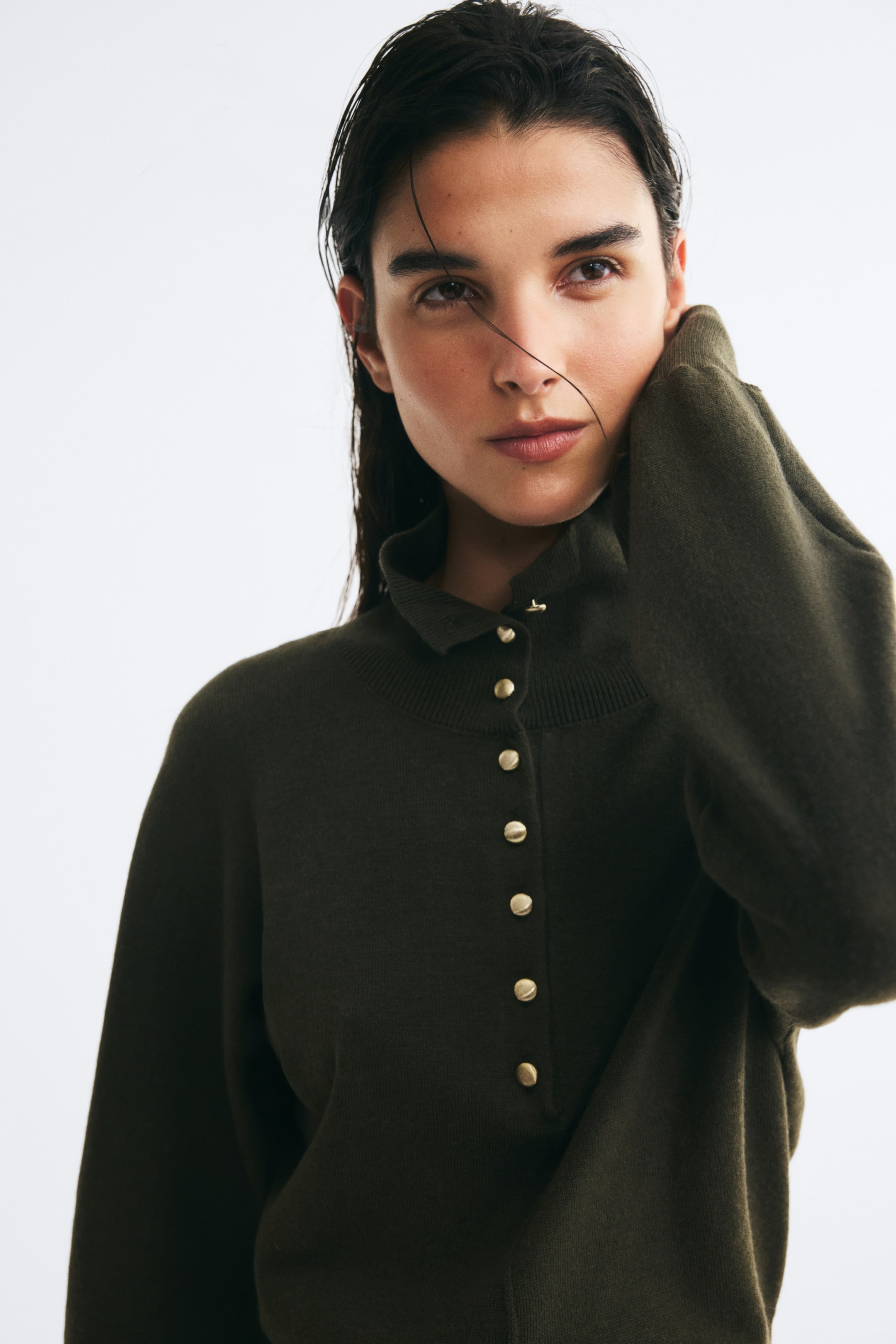Se større billede: En mørkegrøn, fintstrikket sweater med høj hals, ribkrave og en lodret stolpelukning med knapper i guldfarvet finish bæres af modellen. Modellen hviler sin hånd mod siden af halsen og fremhæver garmentets lange ærme.