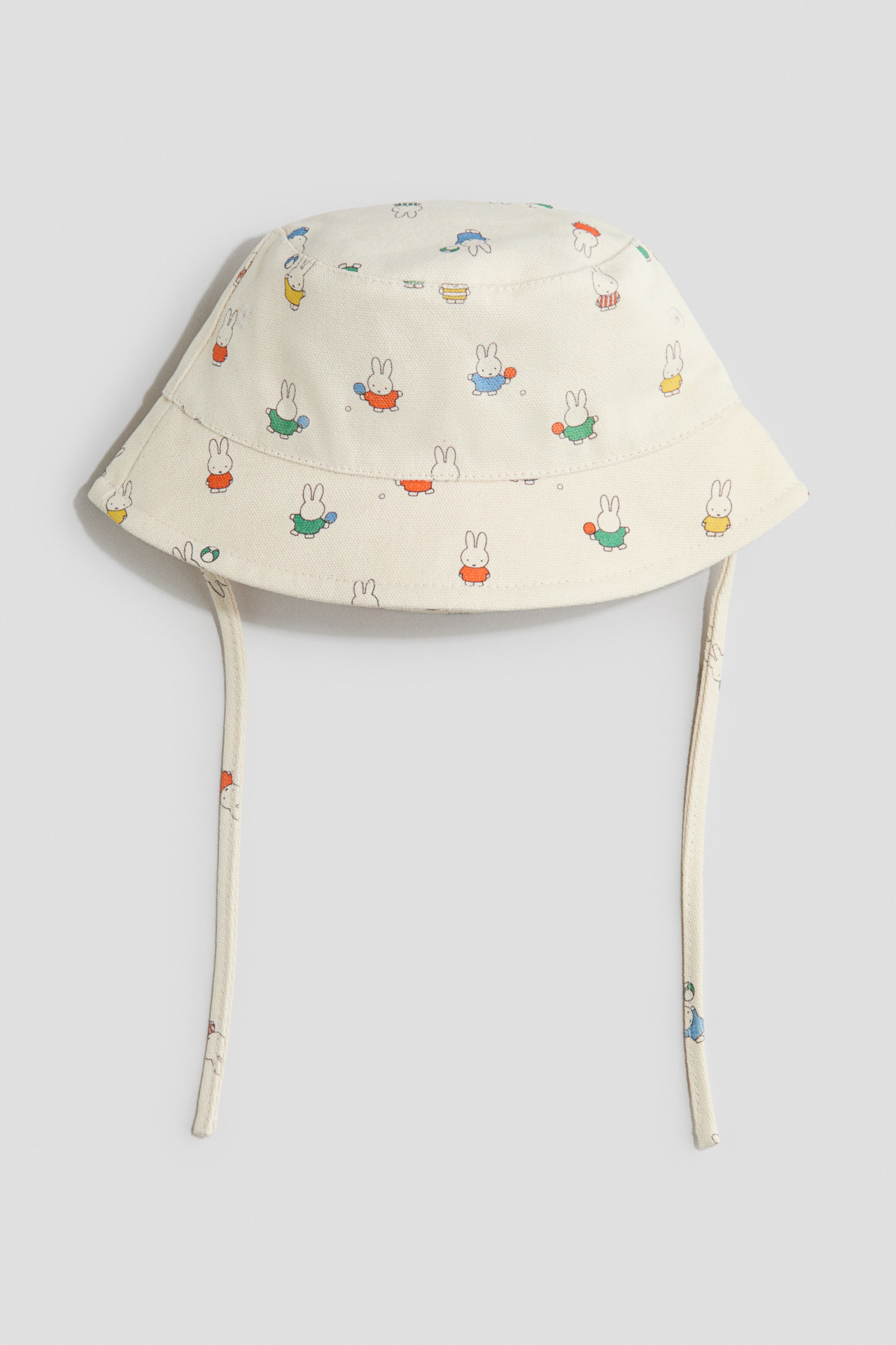 View larger image: A cream cotton canvas bucket hat showcases an all-over print of small, vibrant rabbit characters. Displayed flat, it highlights the front, with a flat crown, a downward-sloping brim, and two matching fabric ties extending from its sides.