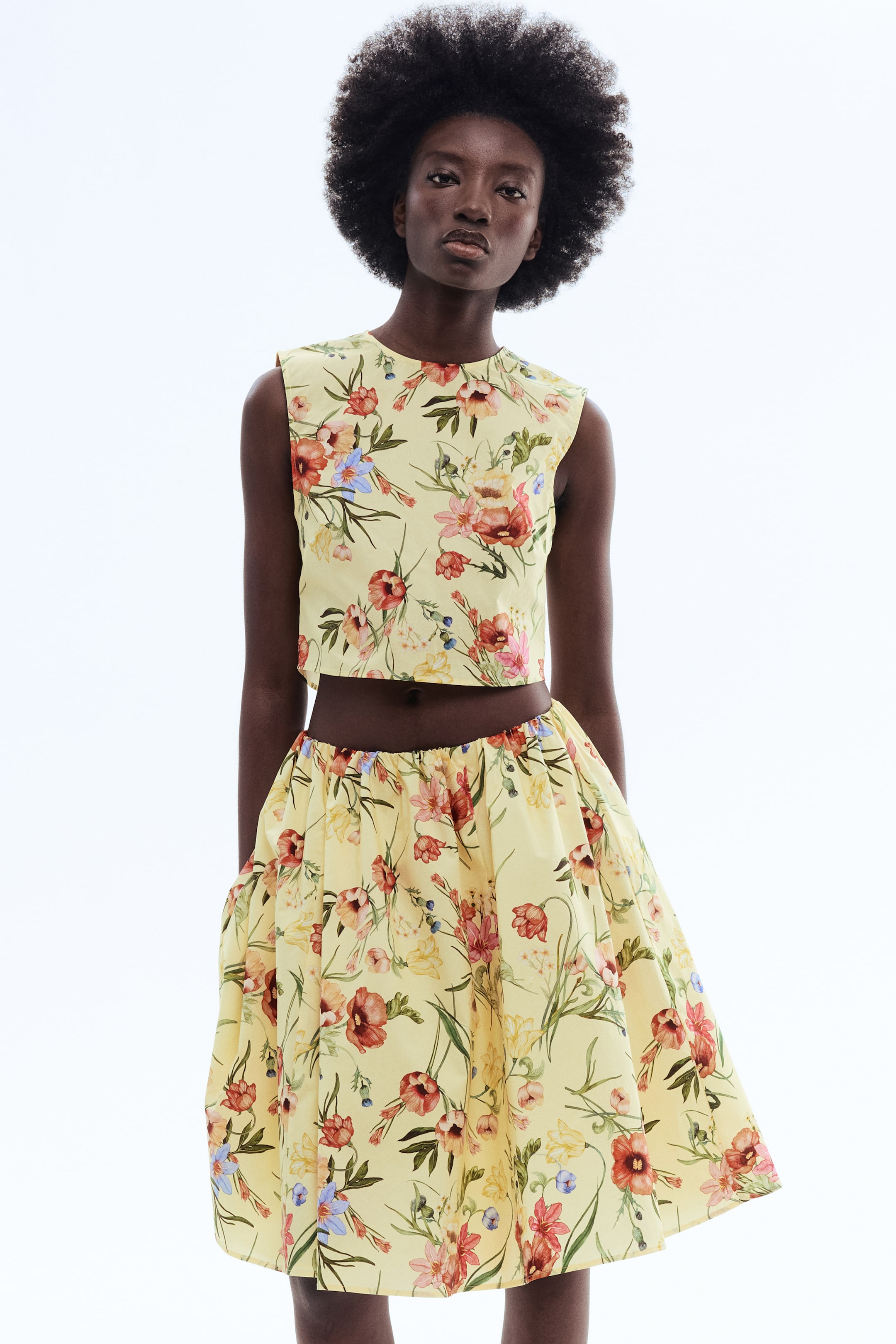 View larger image: A light yellow two-piece set is showcased, featuring a sleeveless crop top with a round neckline and a matching flared skirt with a gathered waist, both adorned with a vibrant floral print.