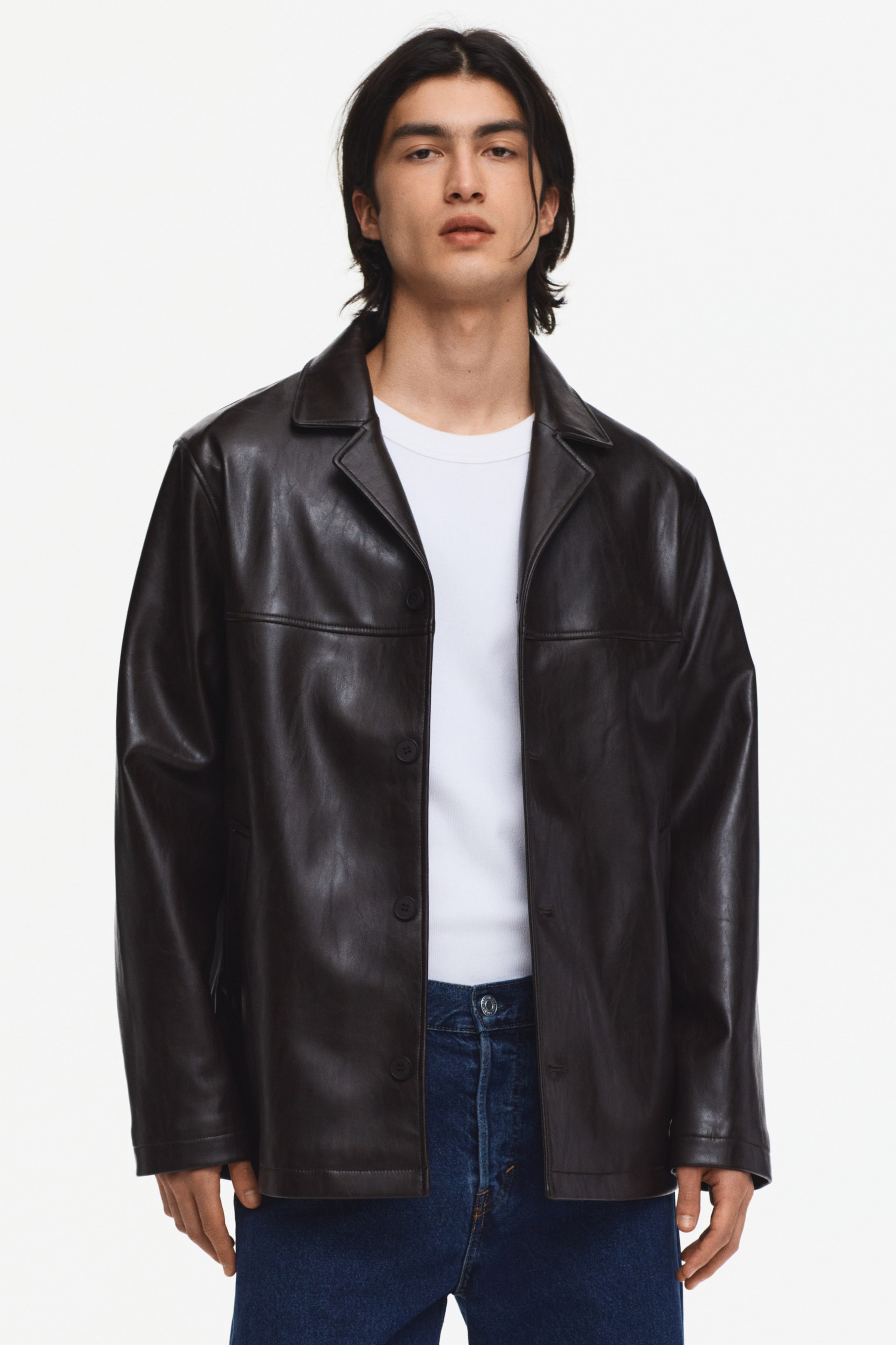 拡大画像を表示: ダークブラウンのフェイクレザージャケットが紹介されています。特徴として、尖った襟、2つの水平な胸の縫い目、そして4つのボタンが見えるボタンフロントの閉じ具があります。白いTシャツと青いジーンズの上にオープンに着用されており、長袖、ストレートヘム、リラックスしたフィット感が際立っています。