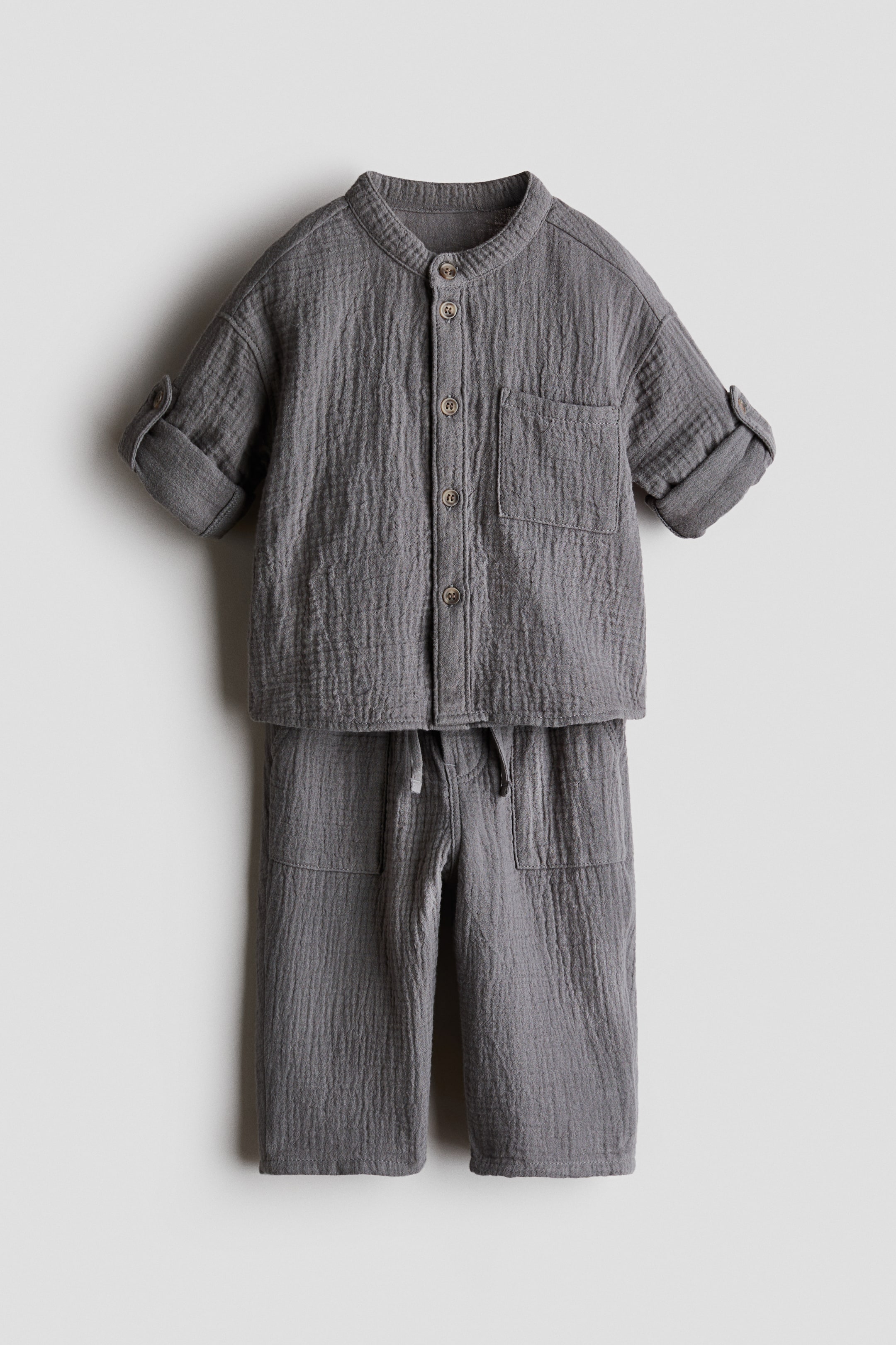 View larger image: "A grey cotton muslin two-piece set is showcased FRONT, featuring a shirt with a band collar, button-down front, open chest pocket, and long sleeves rolled up and secured with tabs and buttons. The matching trousers are highlighted with an elasticated drawstring waist and side pockets."
