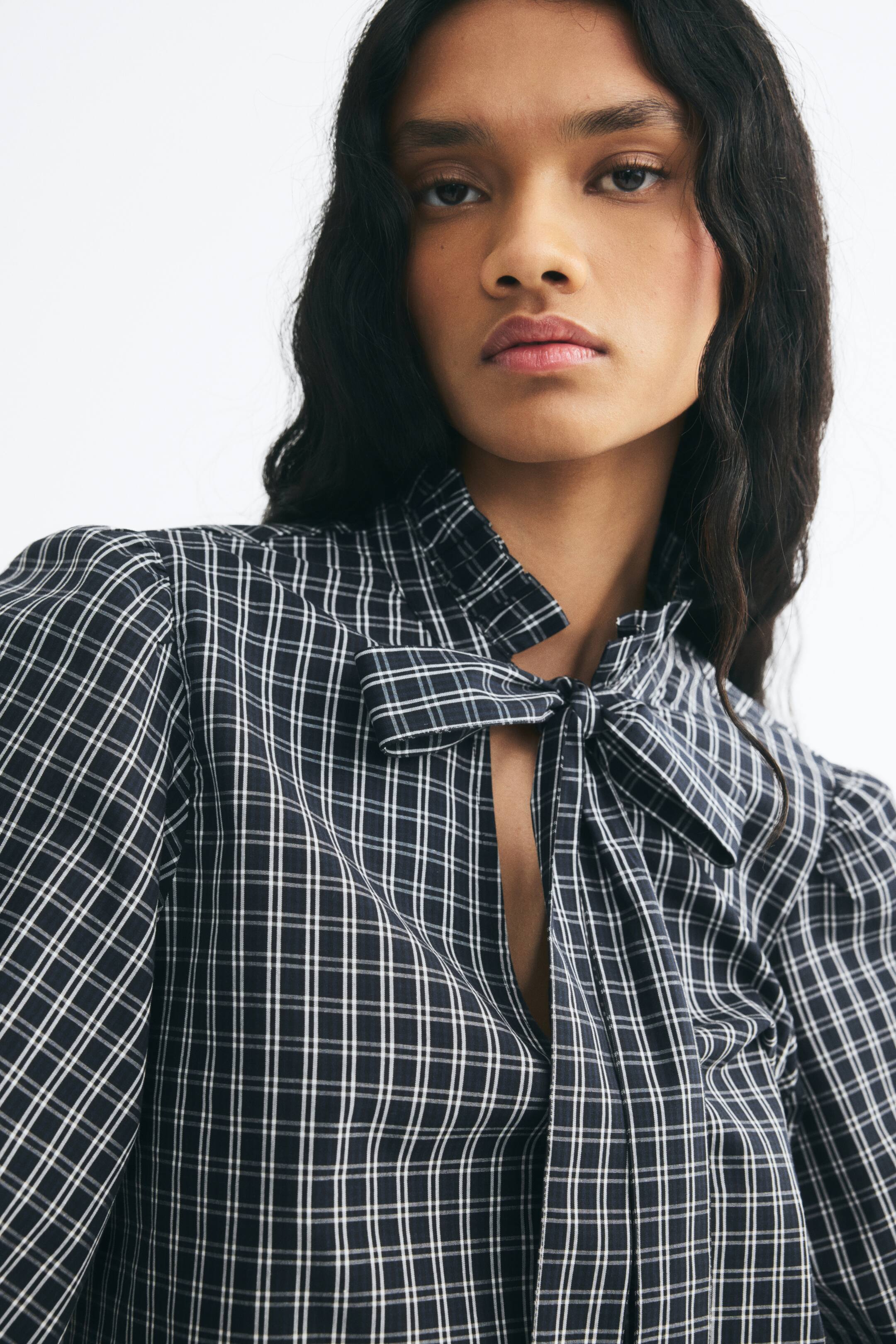 View larger image: A dark, plaid-patterned blouse is being worn by the model, featuring a high collar with frill trim. The blouse has a front keyhole opening tied with a bow, and long sleeves with gathered shoulders.