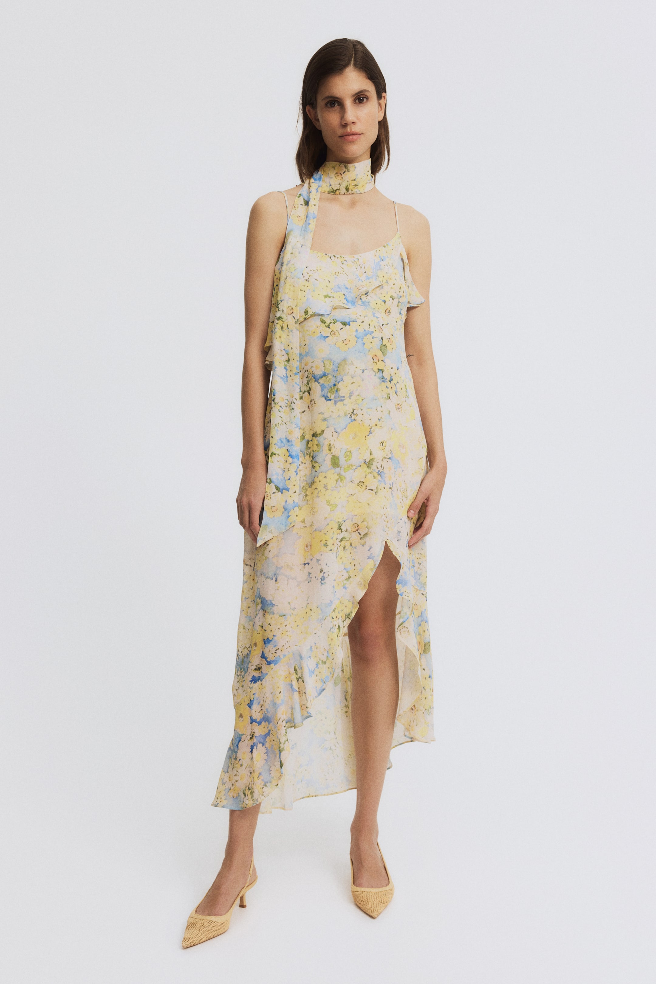 Büyük resmi görüntüle: Sarı ve mavi çiçek desenli midi elbise, spagetti askılar, fırfırlı bir üst katman ve yüksek yırtmaçlı asimetrik fırfırlı etekle öne çıkıyor. Aynı desenli çiçekli bir eşarp boyuna bağlanmıştır.