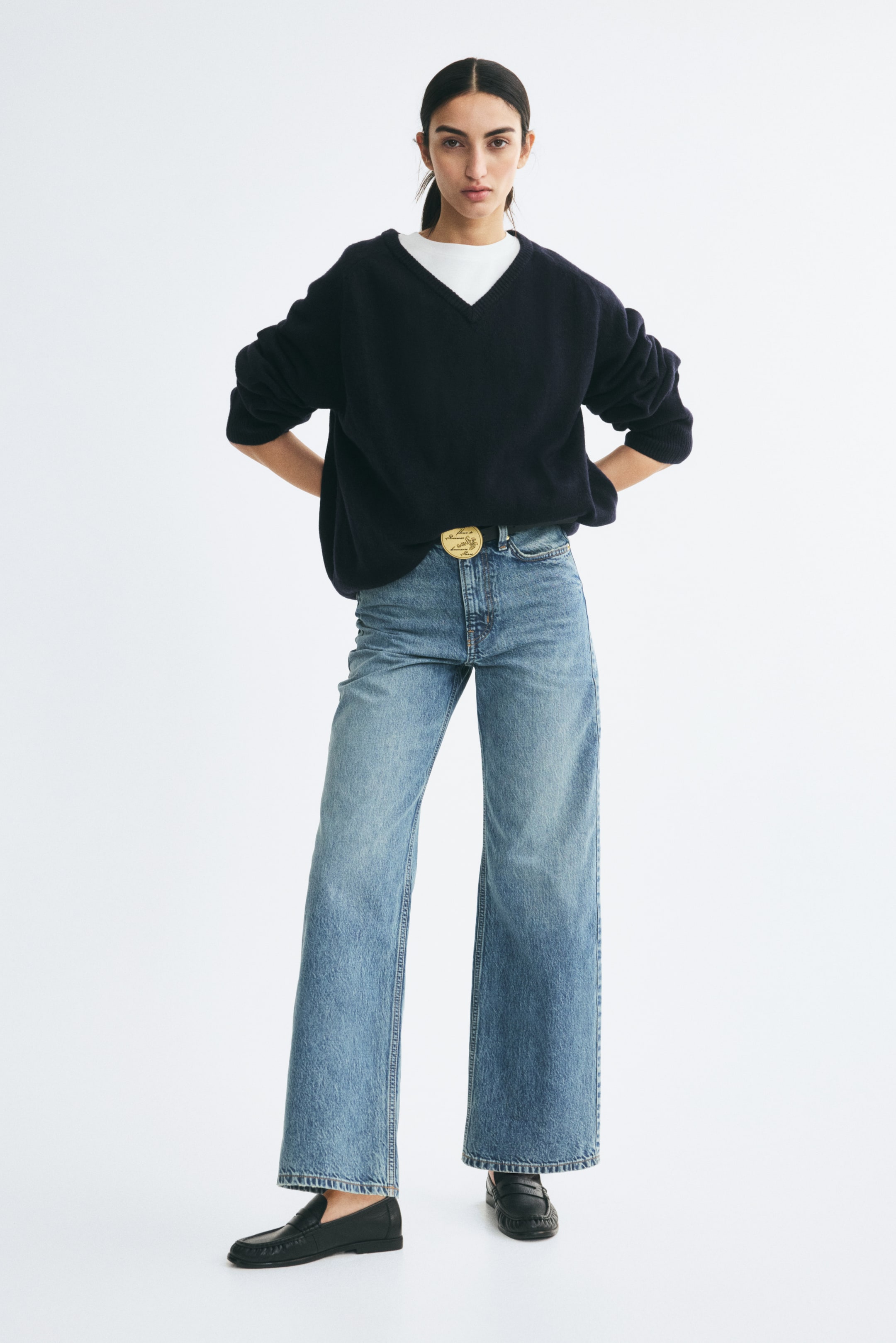 View larger image: High-waisted, straight-leg jeans in a light blue wash are being worn by the model, who is standing facing forward. The jeans feature a relaxed fit through the leg and are styled with a black belt featuring a large gold buckle, a navy V-neck sweater layered over a white T-shirt, and black loafers.