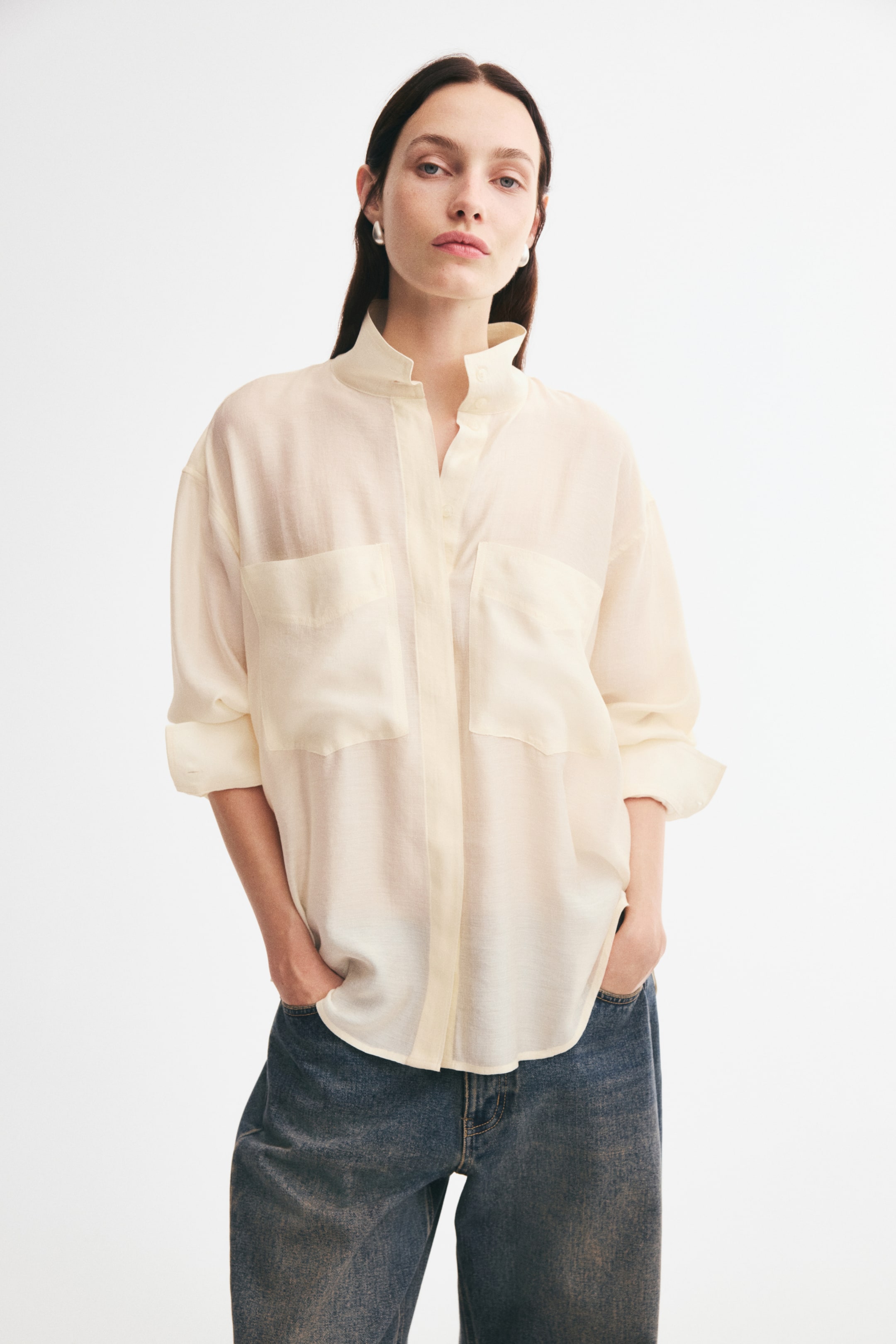 View larger image: An oversized cream-coloured shirt featuring a stand collar, dropped shoulders, and two large patch chest pockets is being worn by the model. The shirt is styled untucked with blue jeans, with the model placing their hands in their front pockets to show the relaxed drape and rounded hem.