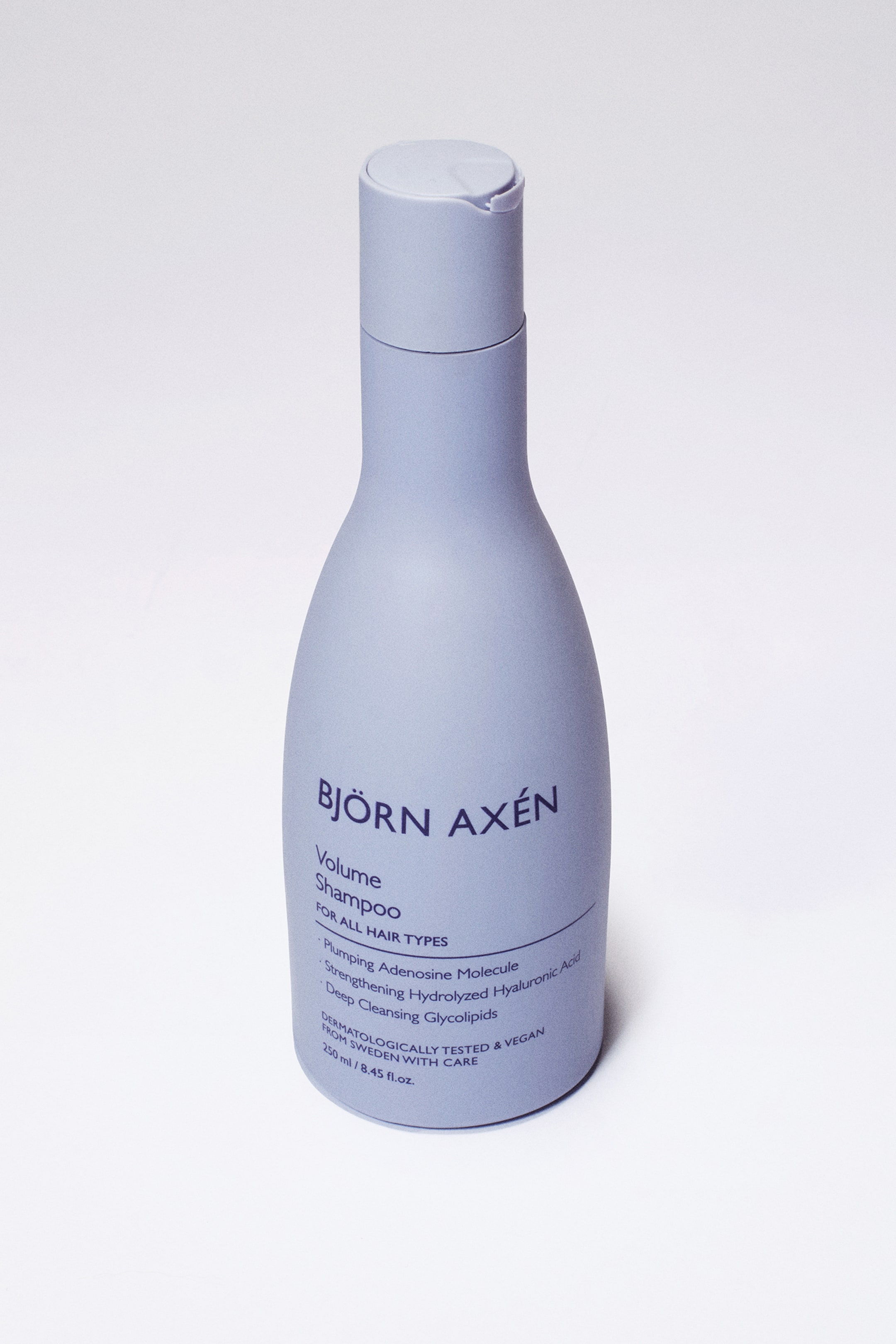 Visa större bild: En ljuslila Björn Axén volymschampo flaska står upprätt, något vinklad åt höger, och visar upp sin frontetikett med "BJÖRN AXÉN Volume Shampoo" i mörka bokstäver. Flaskan har en matt finish och ett matchande flip-top lock.
