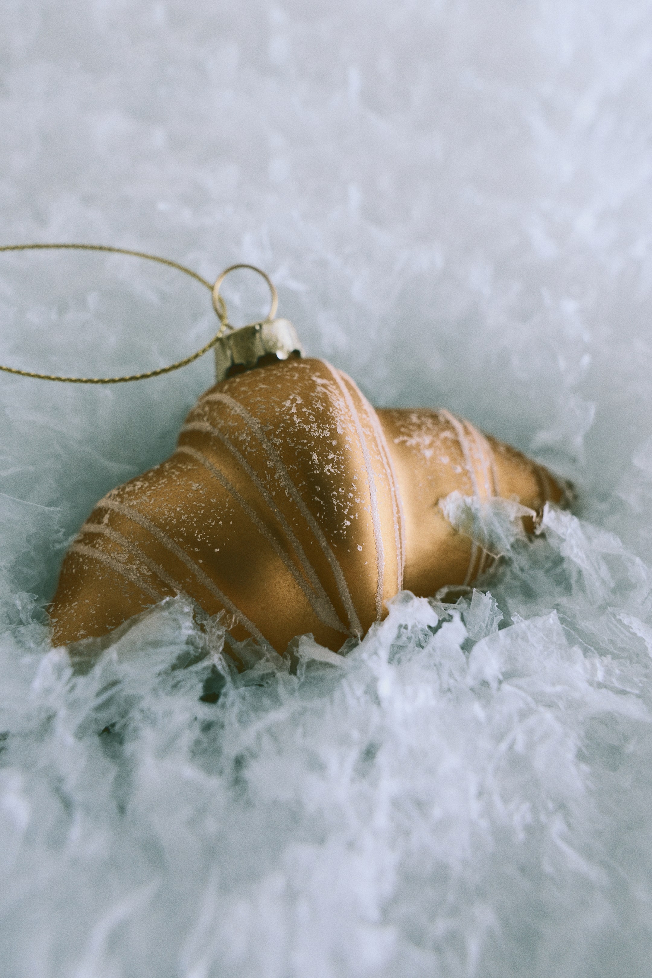 View larger image: A croissant-shaped glass Christmas ornament rests on white textured material, featuring a golden-brown finish with frosted white accents along its curved ridges. A gold metal cap with a thin gold string loop extends from the top of the ornament.