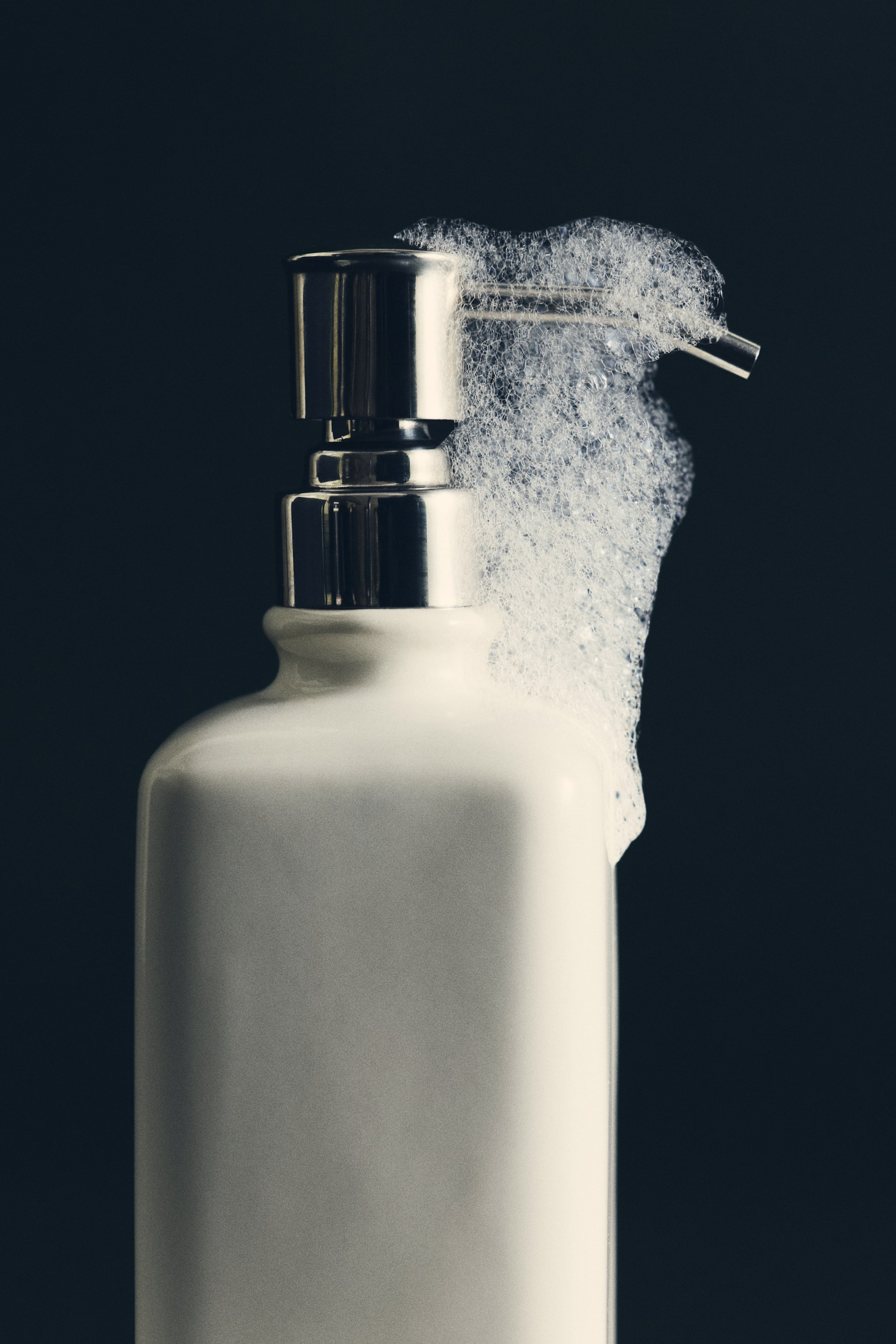 View larger image: A stoneware soap dispenser with a glossy off-white finish is standing upright, featuring a shiny stainless steel pump. White soap foam is dripping from the nozzle down the side of the rectangular body.