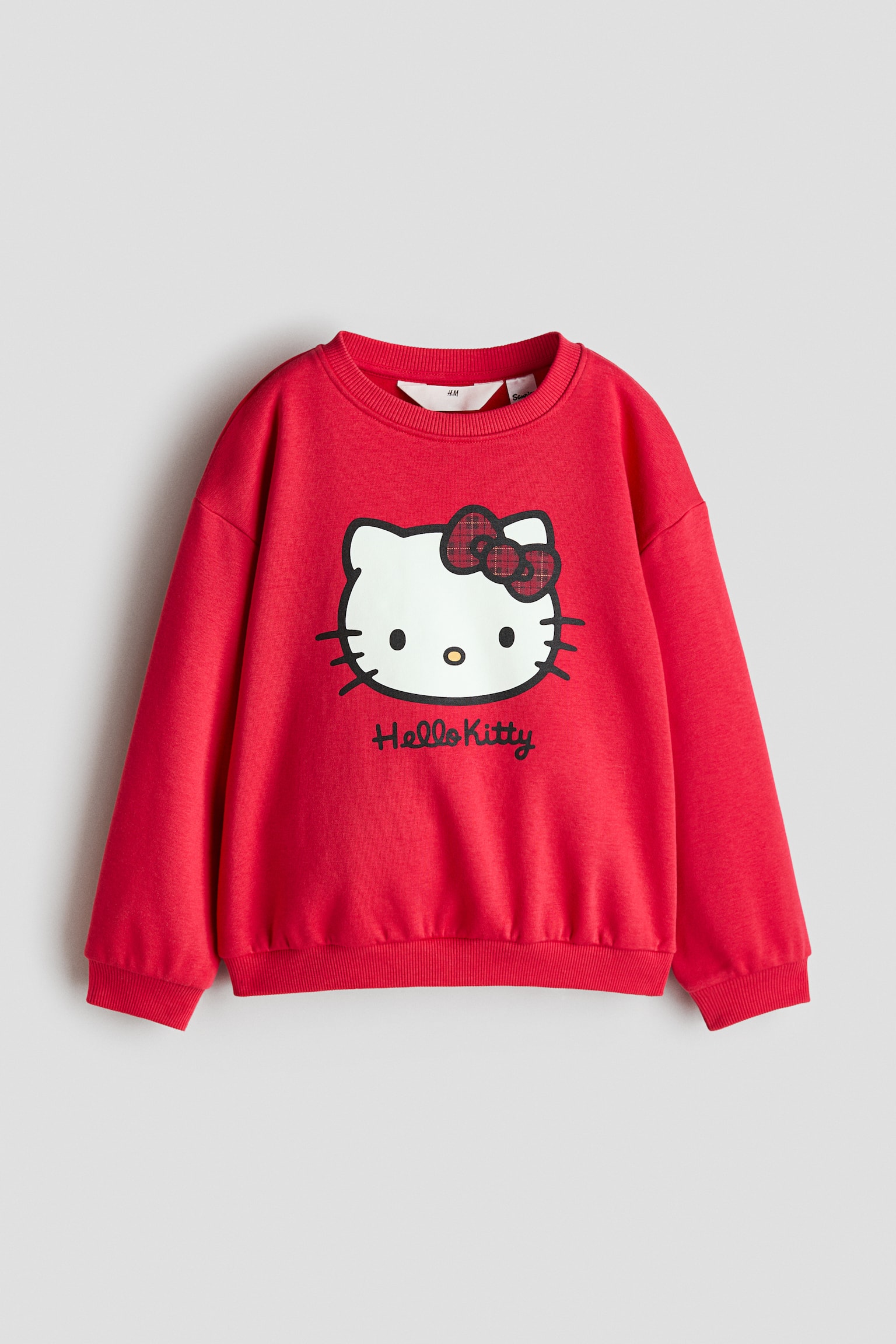 Ein roter Sweatshirt aus Baumwollmischung ist von vorne zu sehen und zeigt auf der Brust einen aufgedruckten Hello Kitty-Print mit einer rot-schwarz karierten Schleife. Das KleidungsstÃ¼ck verfÃ¼gt Ã¼ber einen runden Rippkragen, Ã¼berschnittene Schultern, lange Ãrmel mit RippbÃ¼ndchen und einen gerippten Saum.
