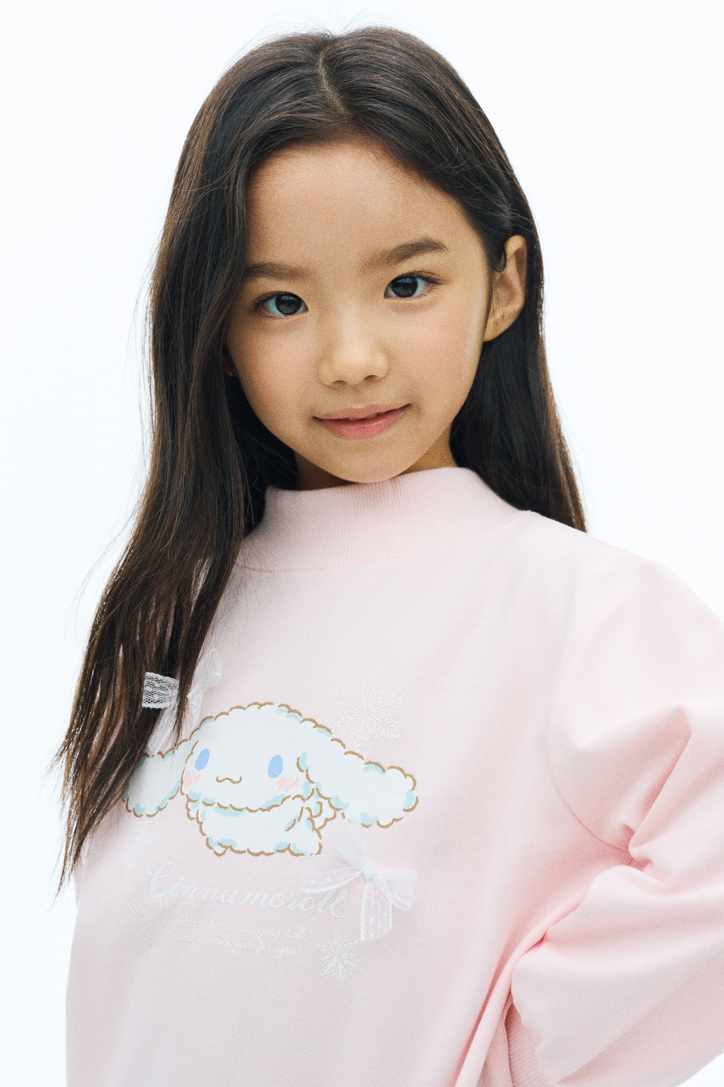 A light pink sweatshirt, shown from the front, displays a printed graphic of the character Cinnamoroll adorned with two small white mesh bows on the chest. The garment features a ribbed mock neck, dropped shoulders, long sleeves with ribbed cuffs, and a ribbed hem.