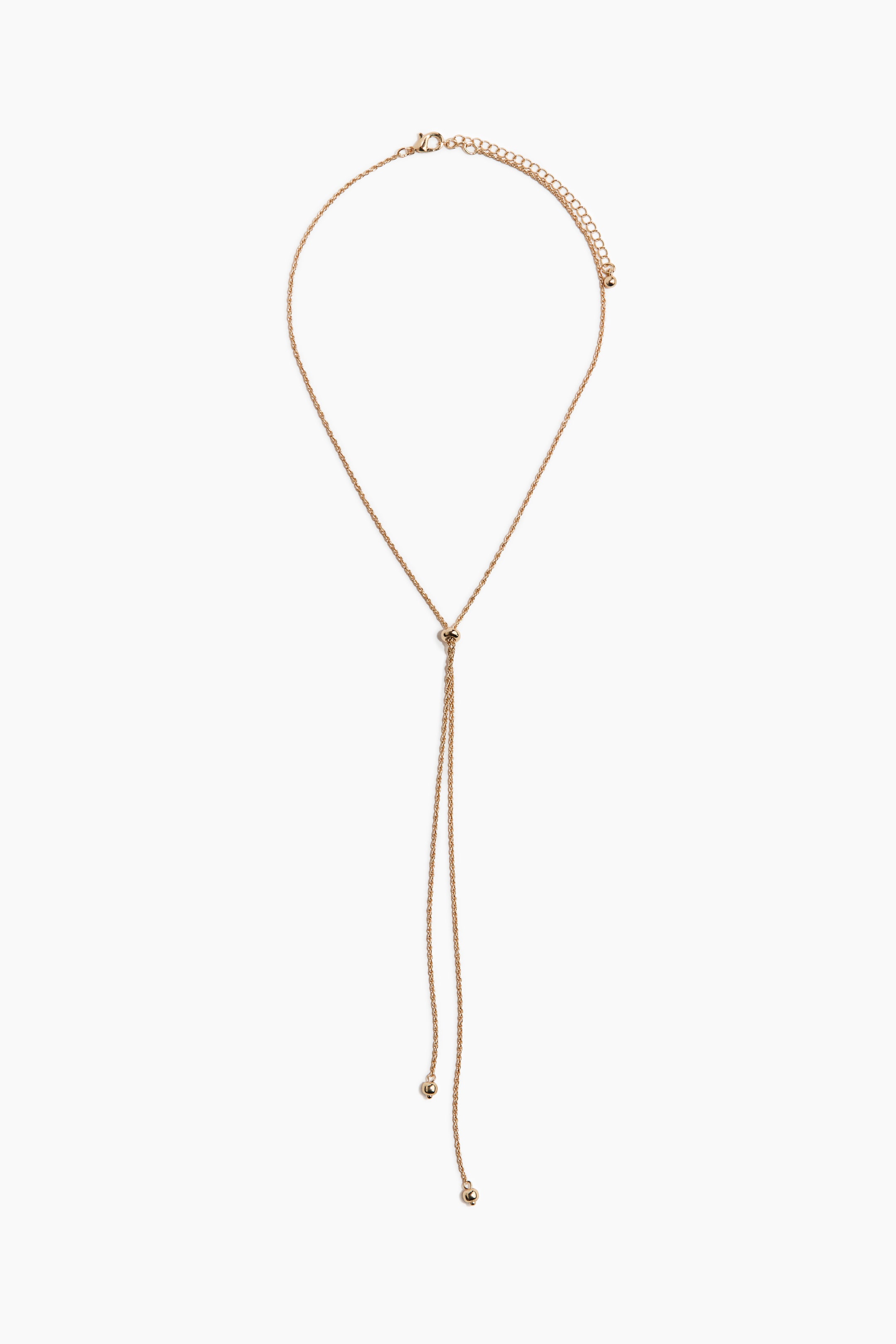 View larger image: The gold-toned metal necklace features a thin chain with an adjustable clasp at the back. It showcases a lariat design at the front, with two dangling chains extending from a small central knot, each ending in a spherical bead.