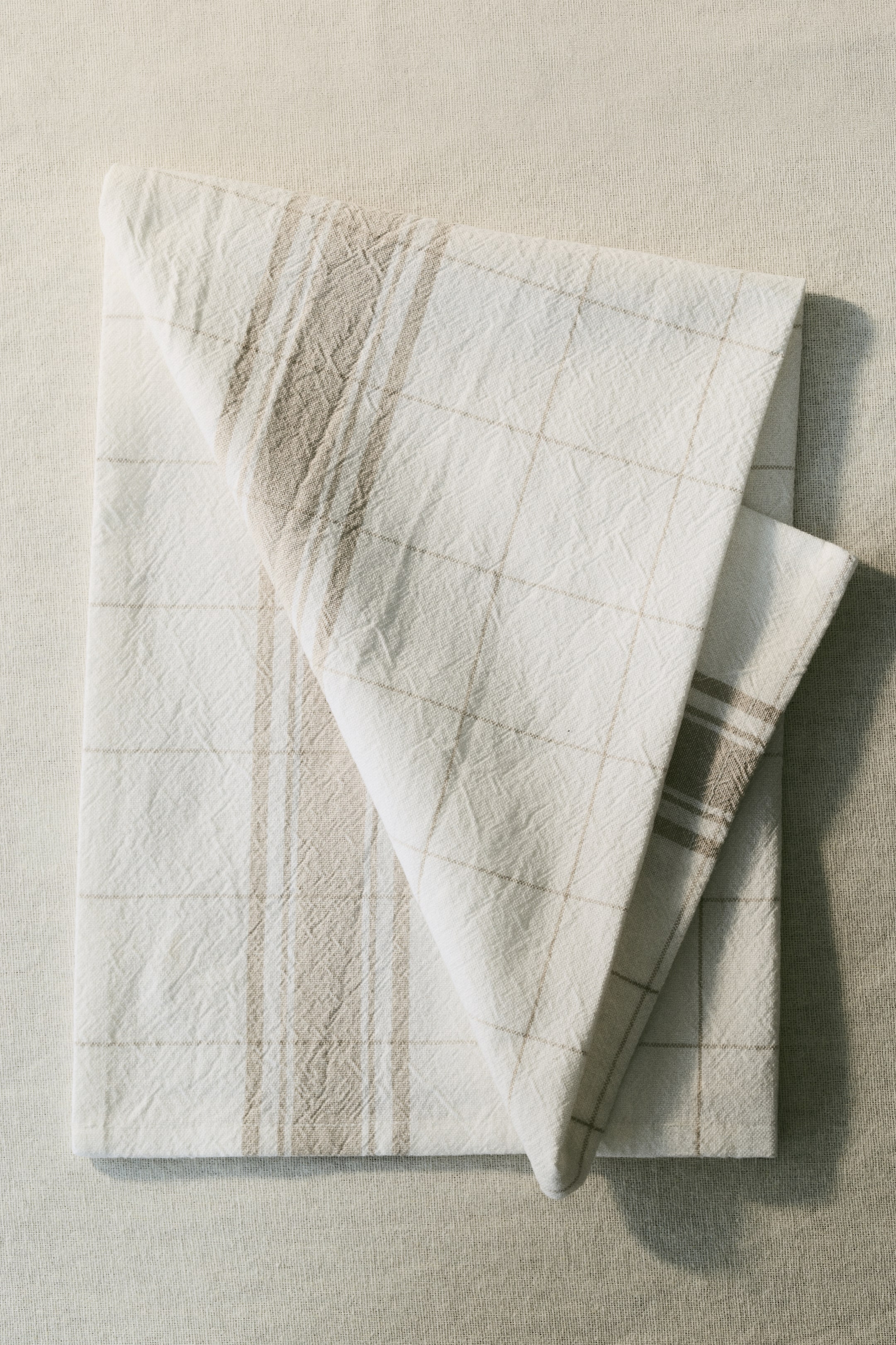 View larger image: A set of three cotton tea towels is shown from the front, folded and stacked. The top towel is displaying an off-white colour with a light brown grid pattern. The towel underneath is revealing an off-white background with light brown vertical stripes.