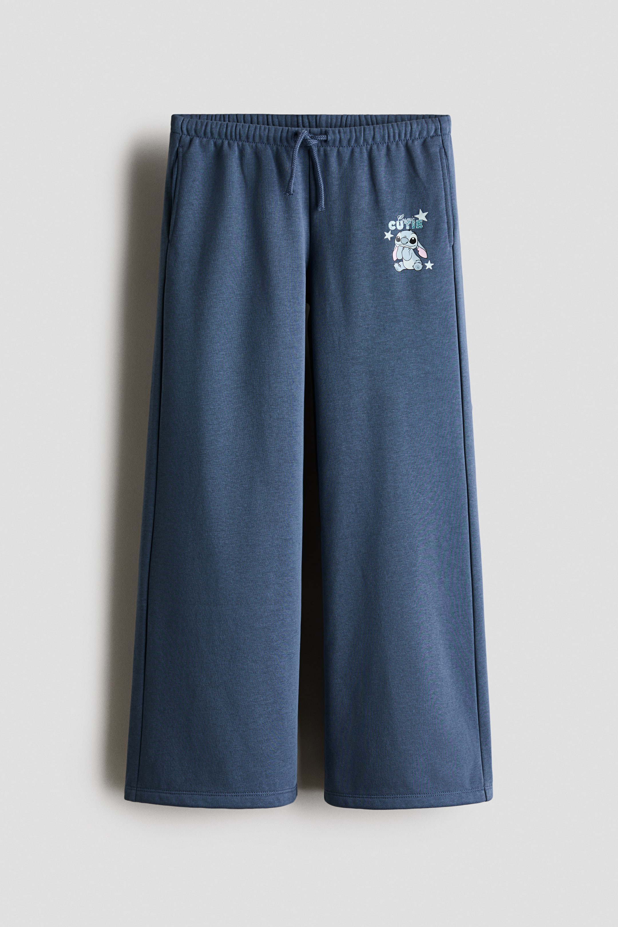 View larger image: Heather blue wide-leg sweatpants featuring an elasticated drawstring waistband with visible ties, side pockets, and a graphic of a light blue cartoon character with “CUTIE” text and stars on the upper right leg.