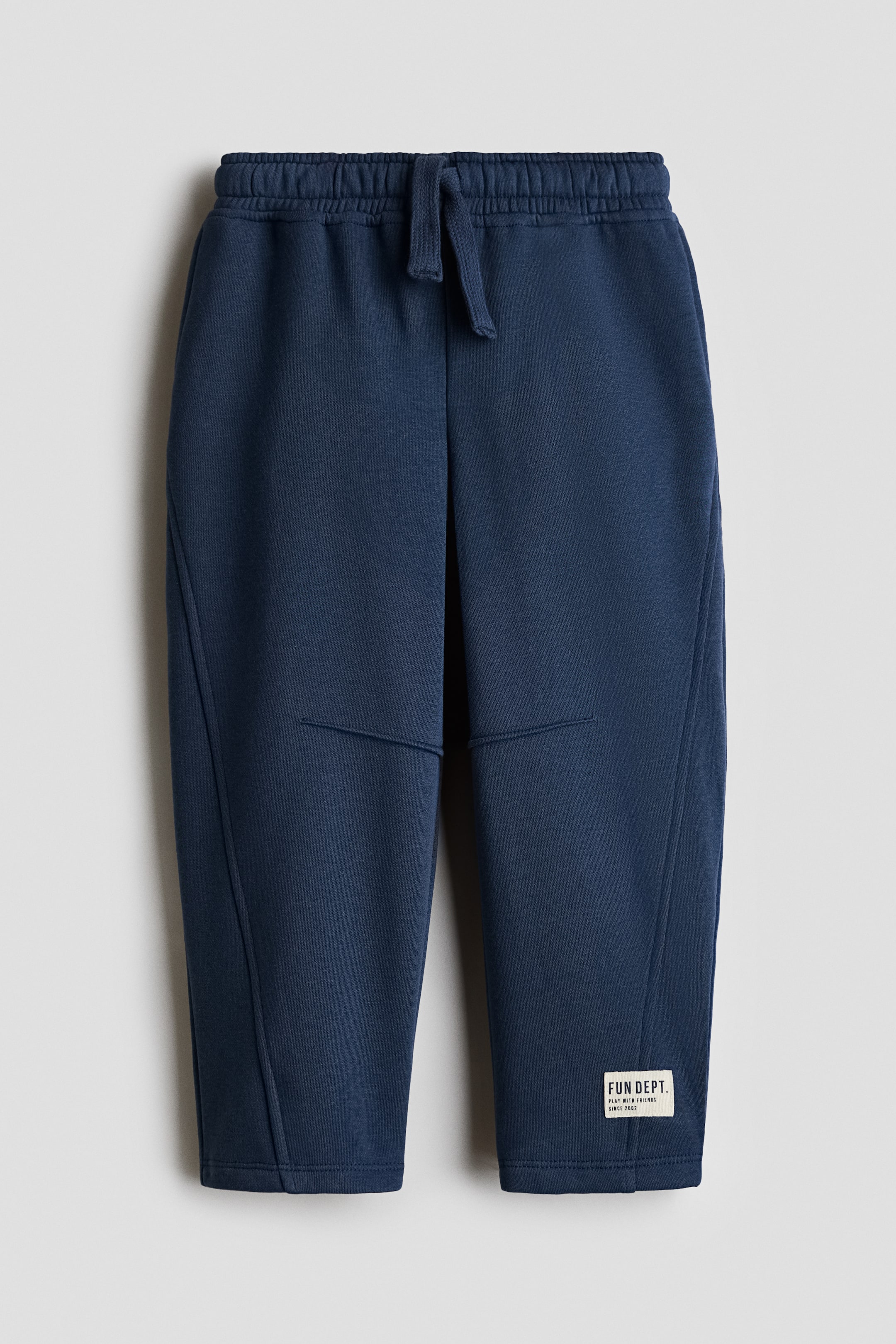 View larger image: The dark blue joggers are made from fleece sweatshirt fabric, featuring an elastic drawstring waist and slightly tapered legs as seen from the front. They are detailed with forward-facing piping down the legs, horizontal seam lines across the knees, and a white brand label on the lower left leg.