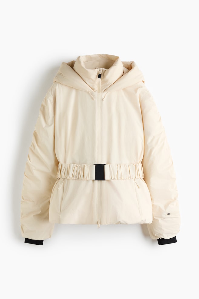 Cette doudoune à capuche couleur crème, vue de face, présente un col montant, une fermeture zippée frontale sur toute la longueur et une ceinture élastiquée intégrée avec une boucle rectangulaire noire. La veste affiche une silhouette matelassée décontractée, avec des manches longues finies par des poignets noirs contrastants. - 1