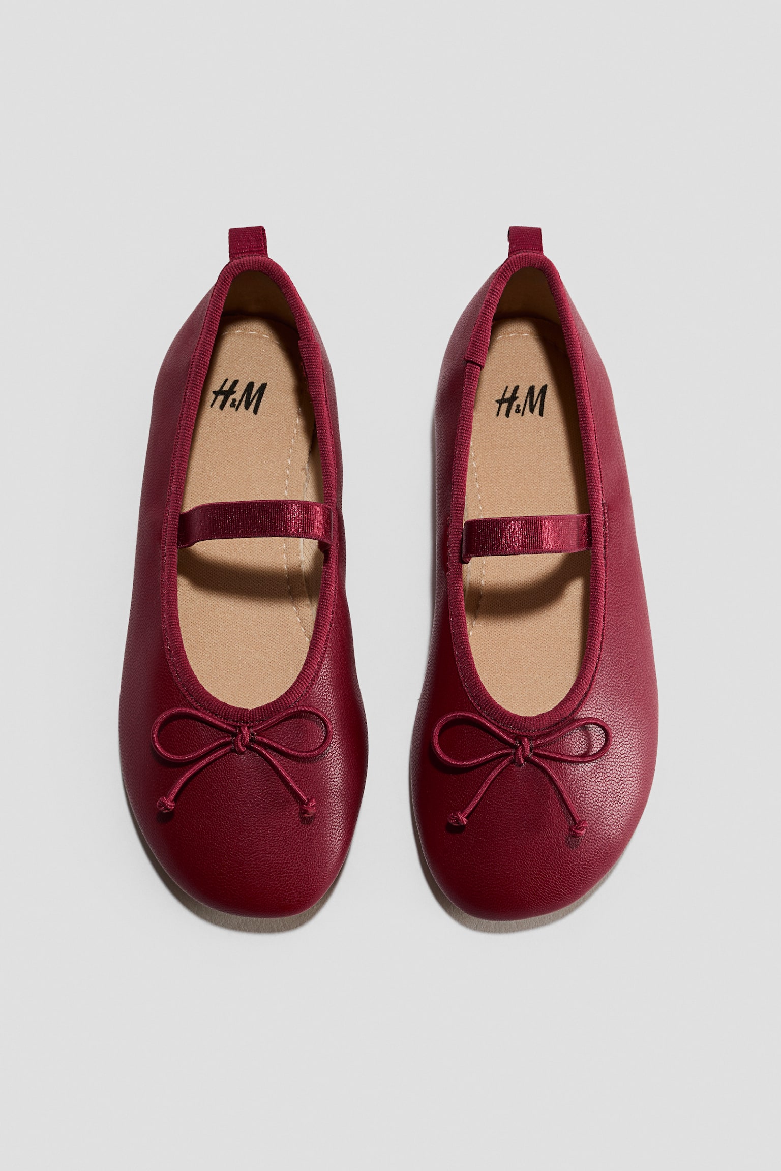 Se muestra un par de bailarinas de color rojo oscuro en vista cenital, con punteras redondeadas y un lazo decorativo en la parte delantera. Los zapatos cuentan con una tira elÃ¡stica sobre el empeine, un ribete de grosgrain a juego alrededor de la abertura y una presilla de tiro en el talÃ³n trasero.