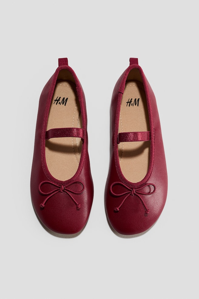 Une paire de ballerines rouge foncé est présentée vue de dessus, avec des bouts arrondis et un nÅud décoratif sur lâavant. Les chaussures présentent une bride élastiquée sur le cou-de-pied, un passepoil en gros-grain assorti autour de lâouverture et une tirette à lâarrière du talon. - 1