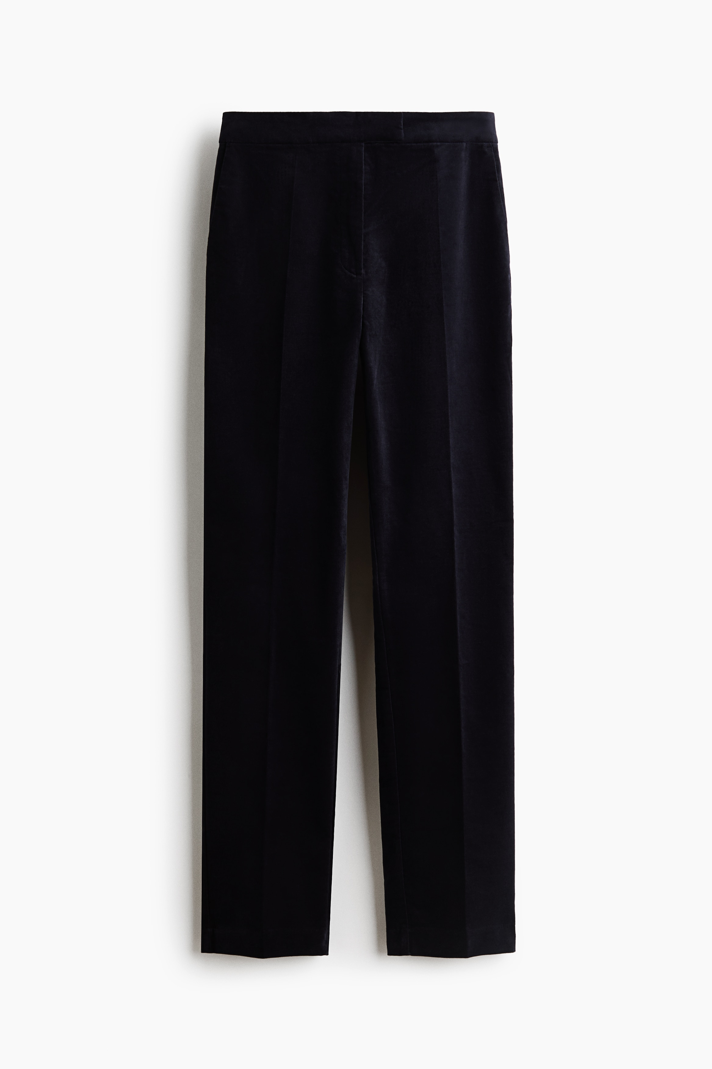 Fitted black velvet pants featuring slim legs with front creases are being worn by the model. The pants are styled with a cream-colored crewneck sweater and black leather flat shoes.
