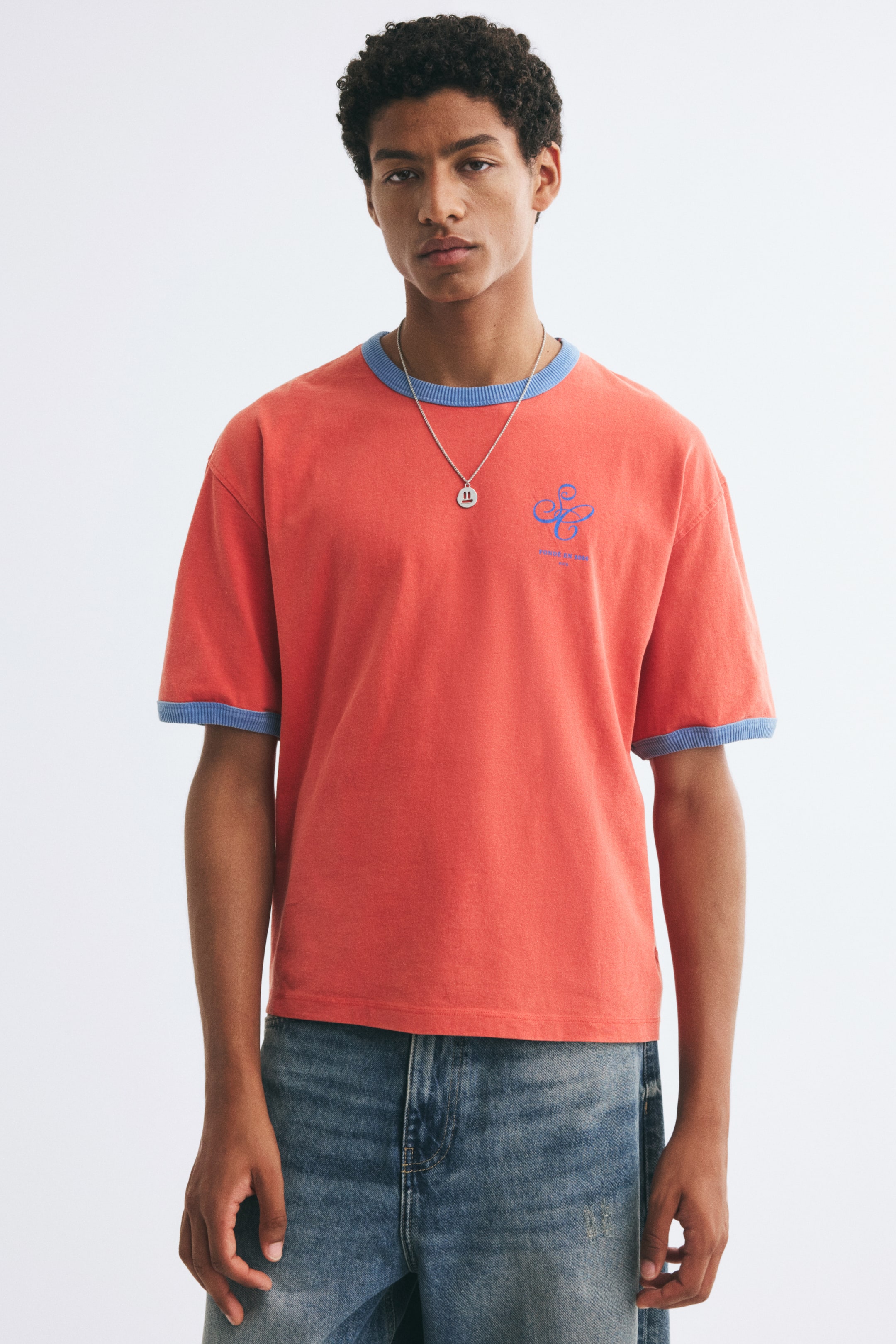 Größeres Bild anzeigen: Ein locker geschnittenes T-Shirt in Korallenfarbe mit kontrastierenden blauen Rippbündchen am Rundhalsausschnitt und an den Ärmelabschlüssen wird von dem Model getragen. Das T-Shirt verfügt über einen kleinen blauen Druck auf der Brust und ist mit blauen Denim-Jeans und einer silbernen Anhängerkette kombiniert.