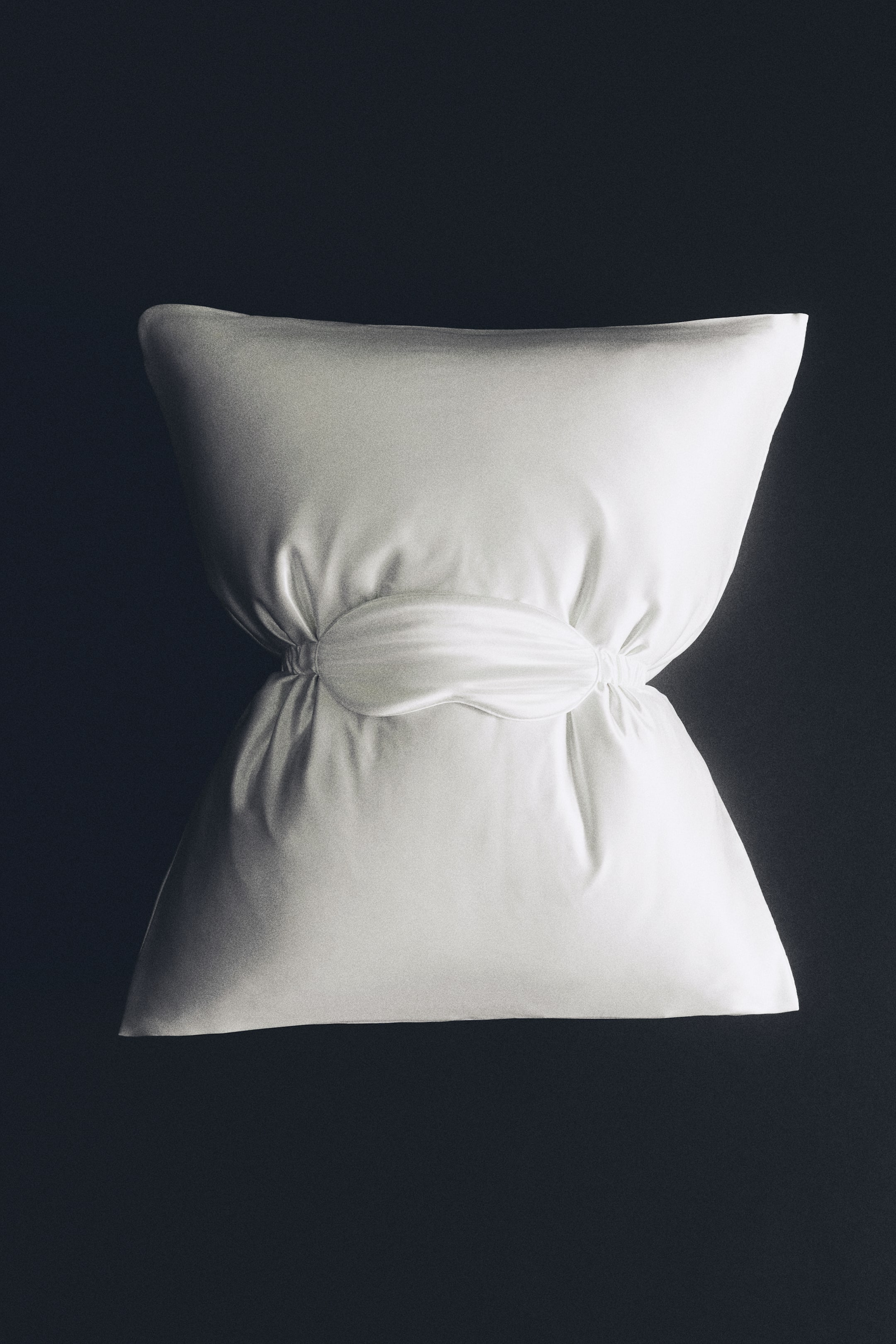View larger image: A white silk pillowcase is cinched at the centre by a matching white silk sleep mask, creating an hourglass shape. The smooth, glossy fabric reflects light and highlights soft gathers around the elastic strap of the sleep mask.