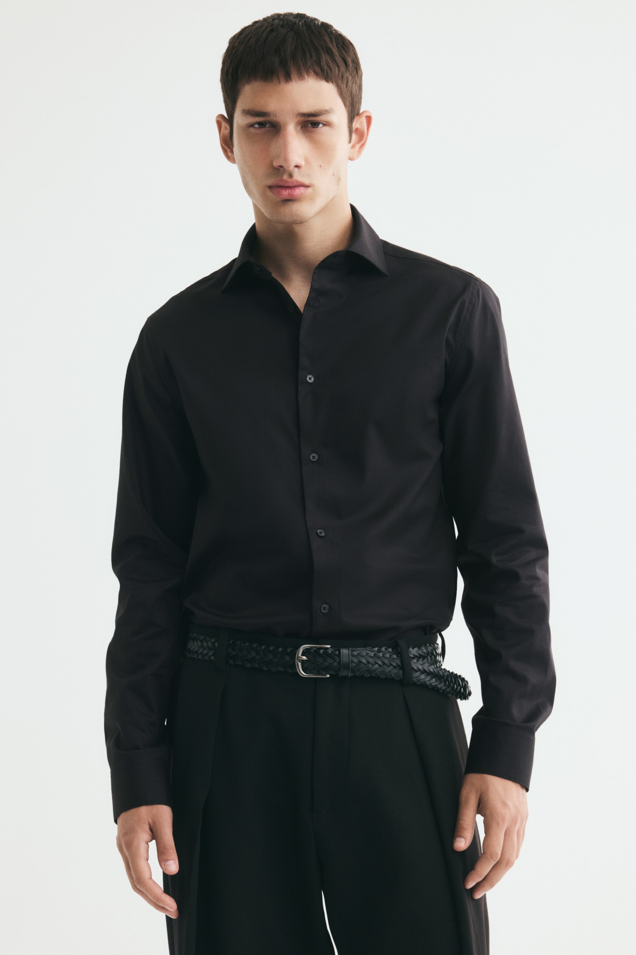 Vezi imaginea mai mare: O cămașă neagră, slim-fit, din bumbac, cu guler tăiat și mâneci lungi, este purtată de model. Cămașa este introdusă în pantaloni negri, asortați cu o curea neagră împletită.