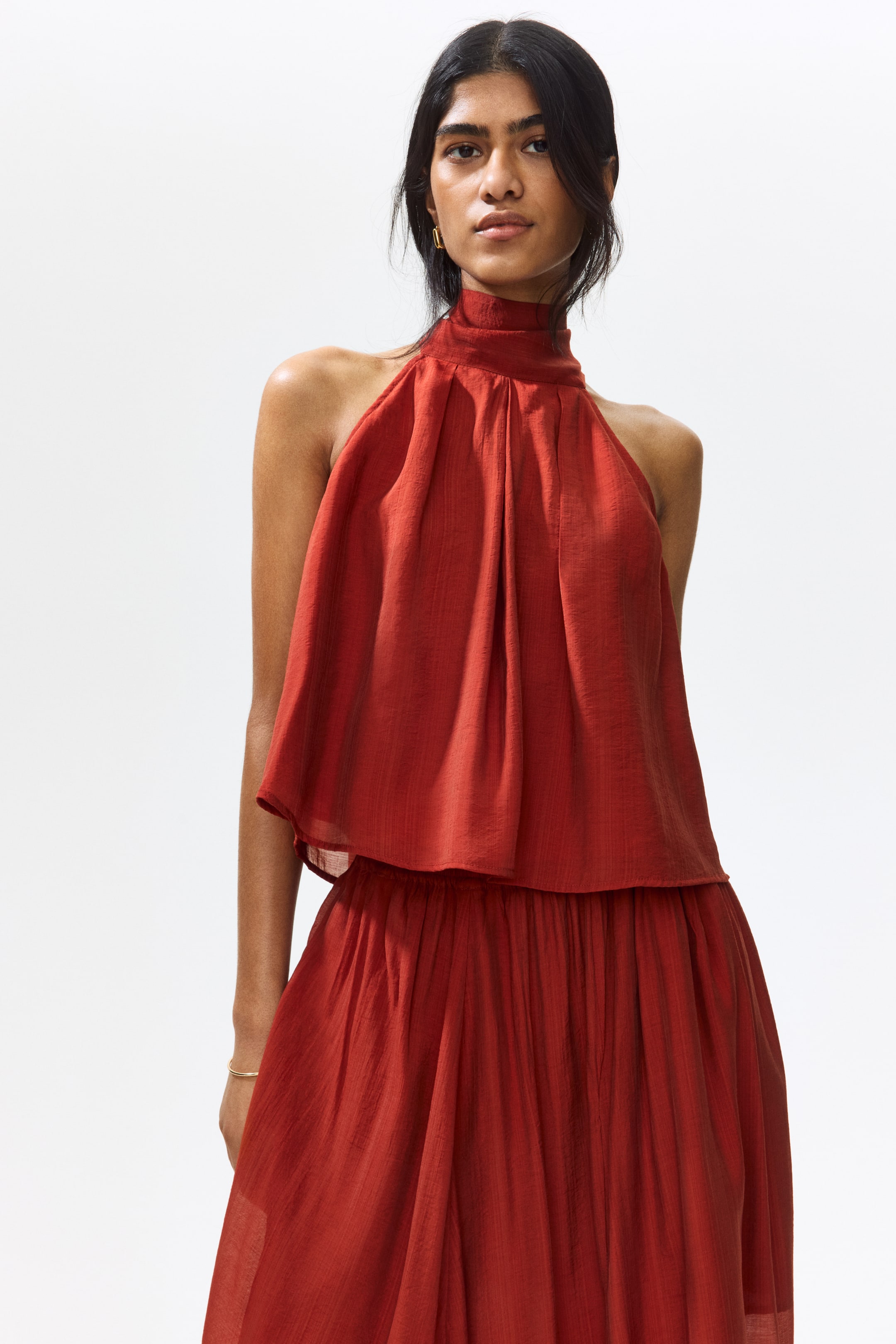 View larger image: The model is showcasing a reddish-brown sleeveless top crafted from woven fabric. It comes with a stand collar and pleats around the neckline, forming a flared silhouette, perfectly paired with a matching skirt.