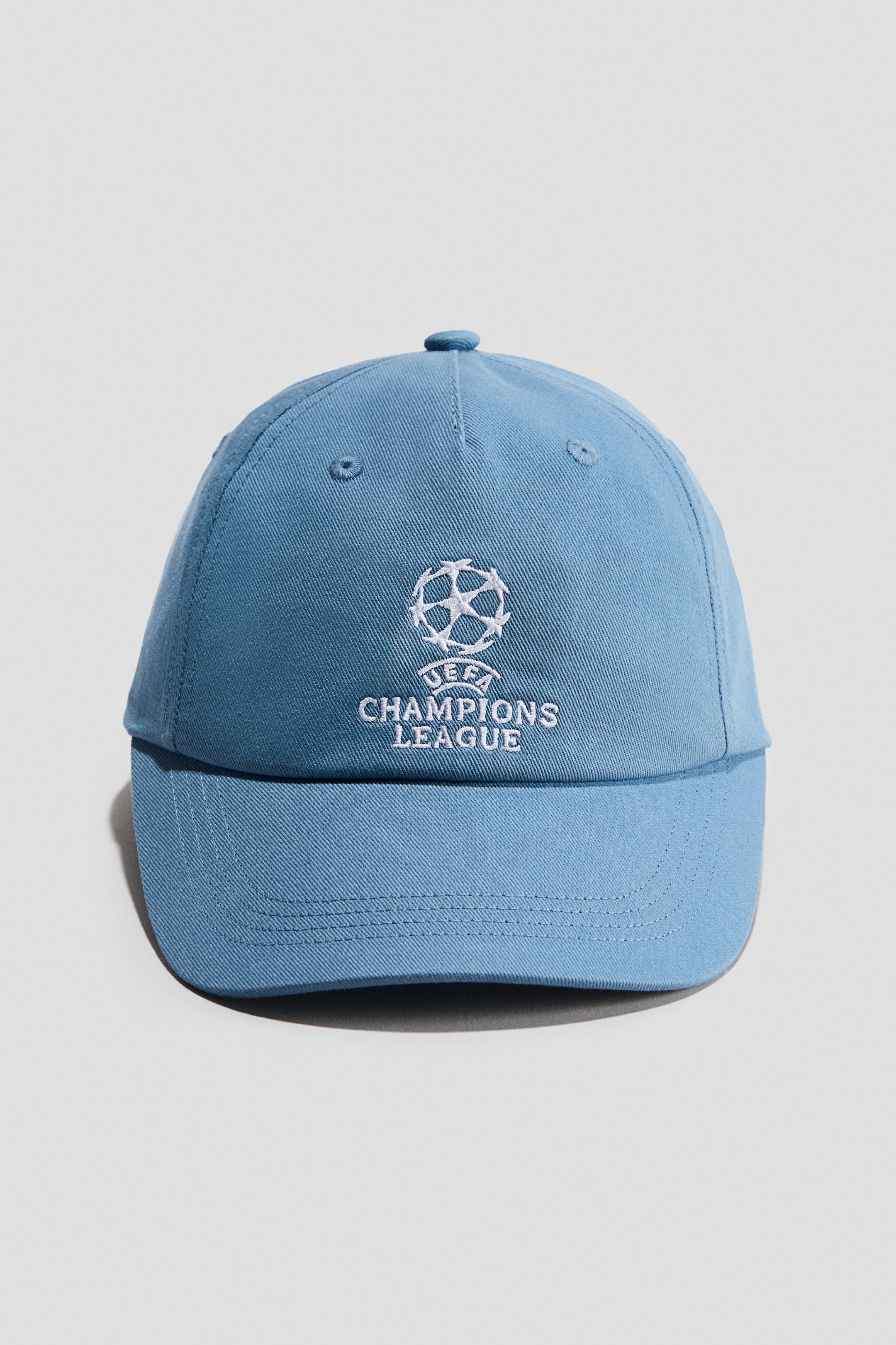 View larger image: A light blue cotton twill cap is facing front, displaying a white embroidered UEFA Champions League logo at the front centre. The cap features a curved peak with tonal stitching, matching embroidered eyelets, and a fabric-covered button on the crown.
