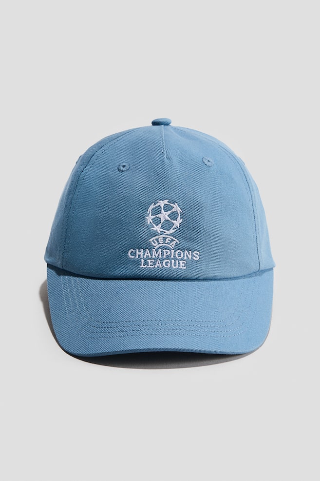 A light blue cotton twill cap is facing forward, displaying a white embroidered UEFA Champions League logo on the front centre. The cap features a curved peak with tonal stitching, matching embroidered eyelets, and a fabric-covered button on the crown. - 1