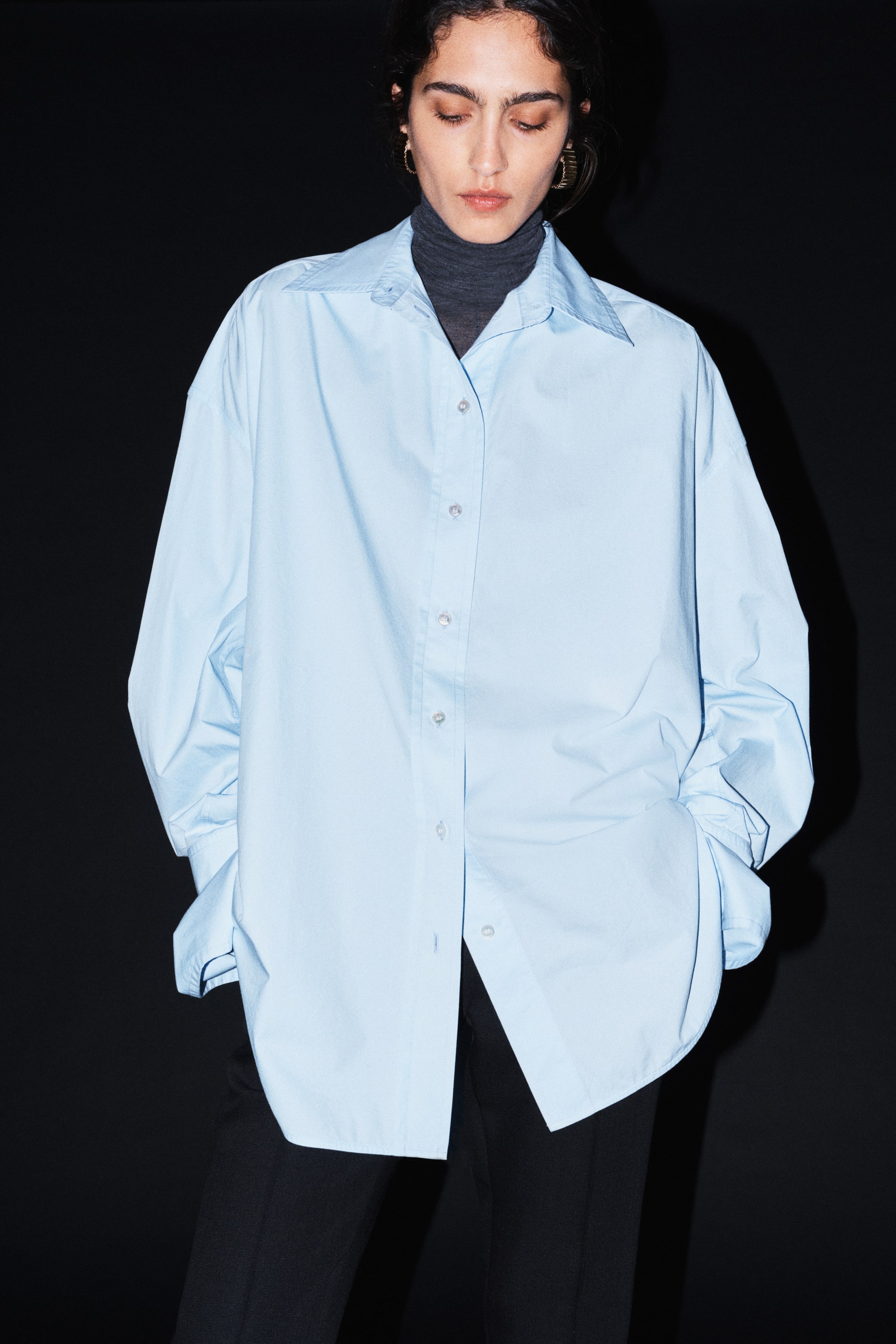 Grösseres Bild ansehen: Ein oversized hellblaues Baumwollhemd mit spitzem Kragen und überschnittenen Schultern wird vom Model über einem dunkelgrauen Rollkragenpullover und schwarzen Hosen getragen. Das langärmelige Hemd verfügt über eine durchgehende Knopfleiste und grosse, umgeschlagene Manschetten, während das Model die Hände in den Taschen hält.