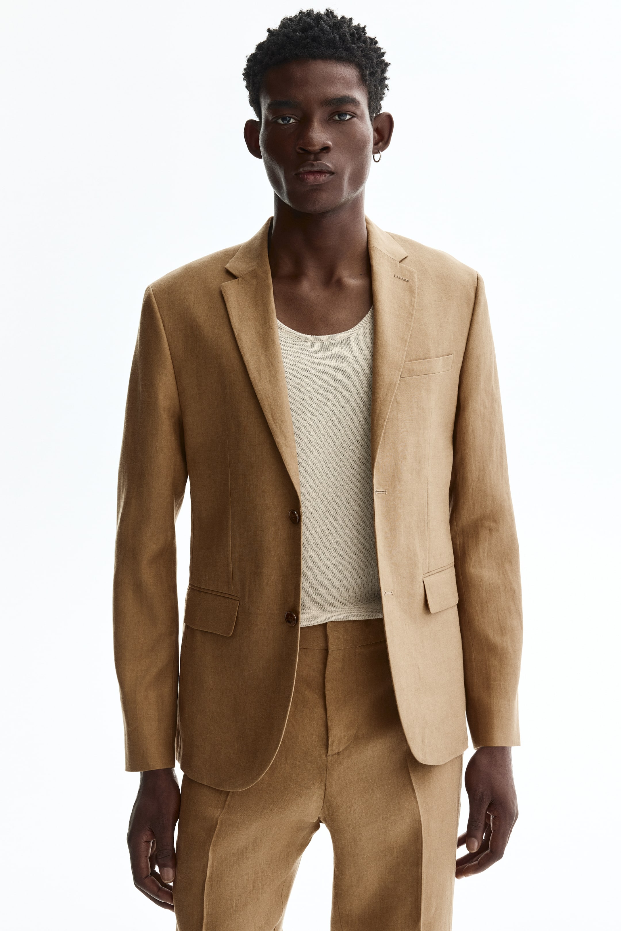View larger image: The model is sporting a single-breasted tan linen jacket, showcasing narrow notch lapels, a decorative buttonhole, a chest pocket, and two flap front pockets. The jacket is secured with one of its two front buttons and is paired with a light knit top and matching linen trousers, achieving a slim, fitted silhouette.